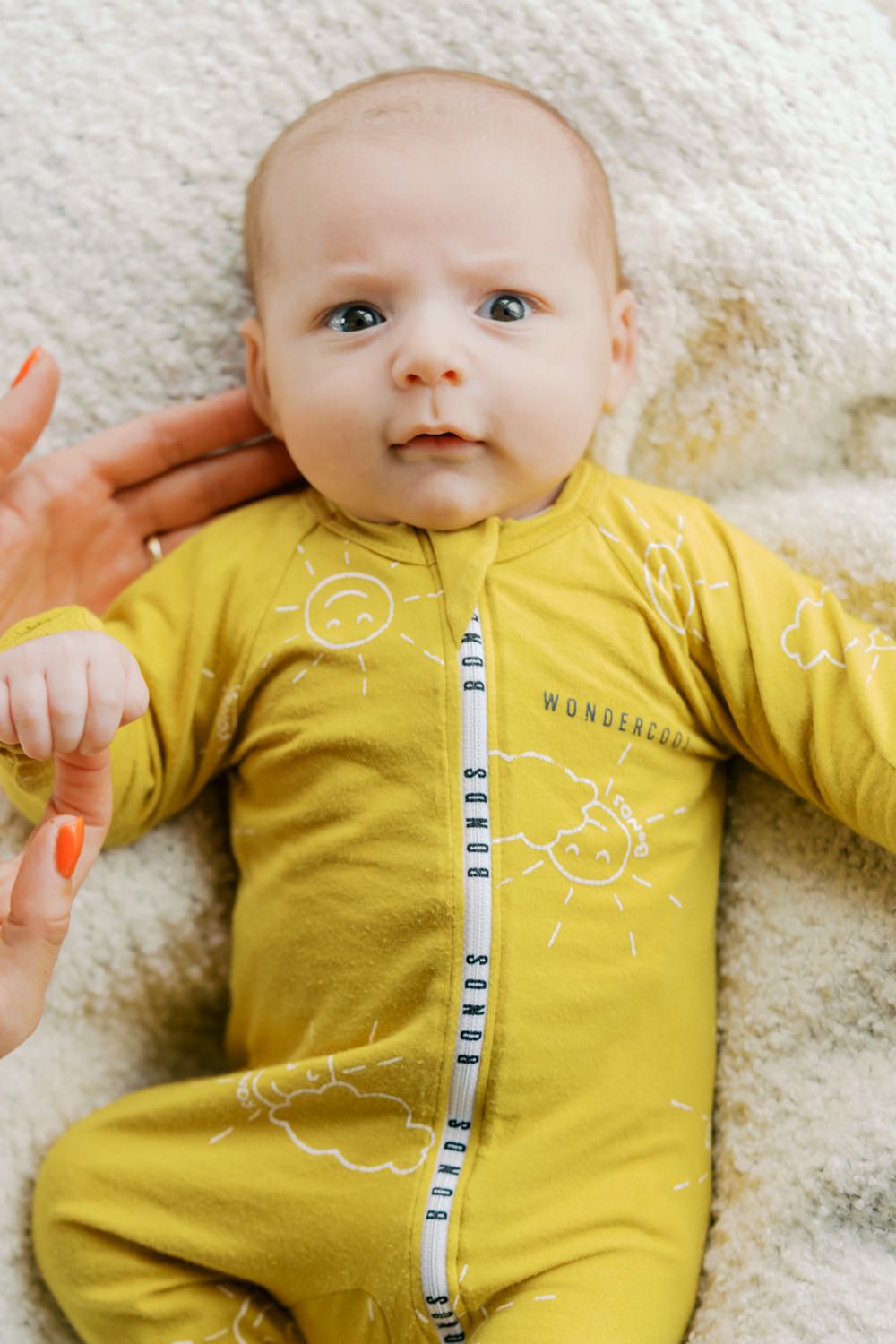 Baby in yellow Wunderkin Co. sunshine print zip-up romper lying on a cream blanket.