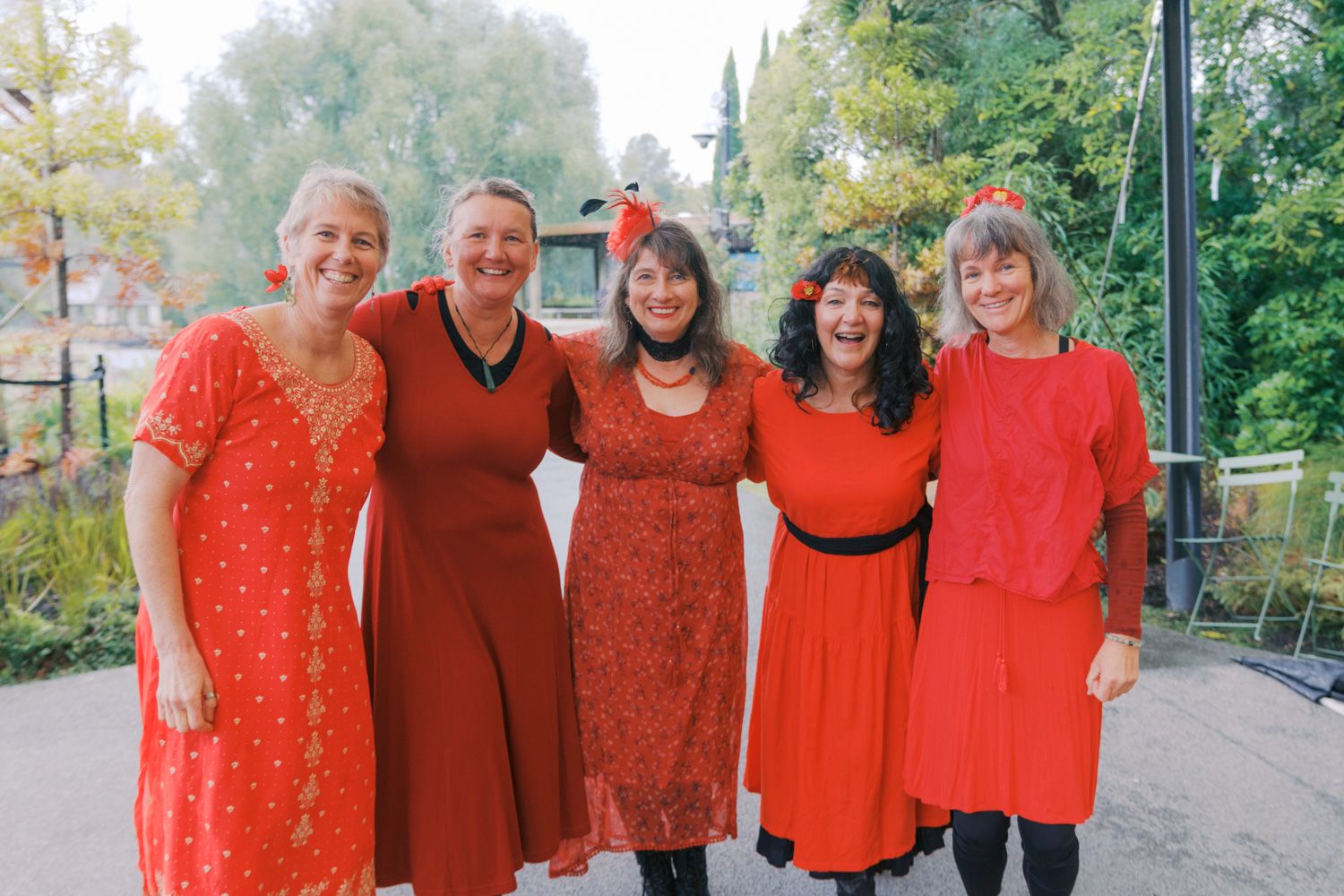 Five women in matching red dresses smiling together outdoors at a garden event.