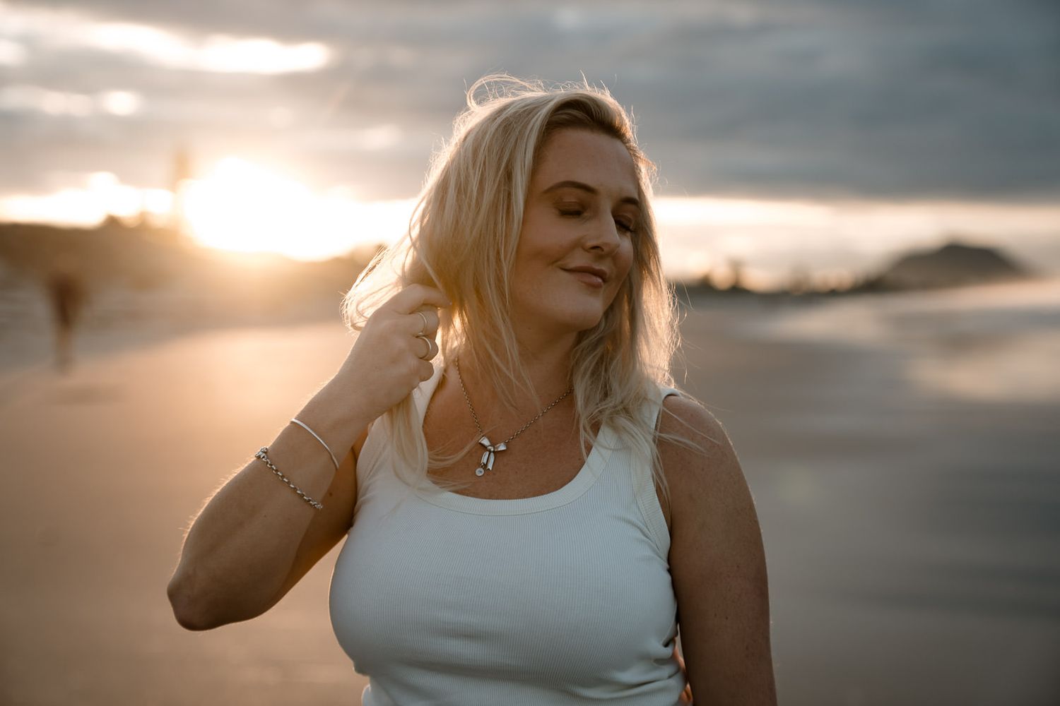 Smiling blonde woman wearing cross necklace on beach during golden hour sunset.