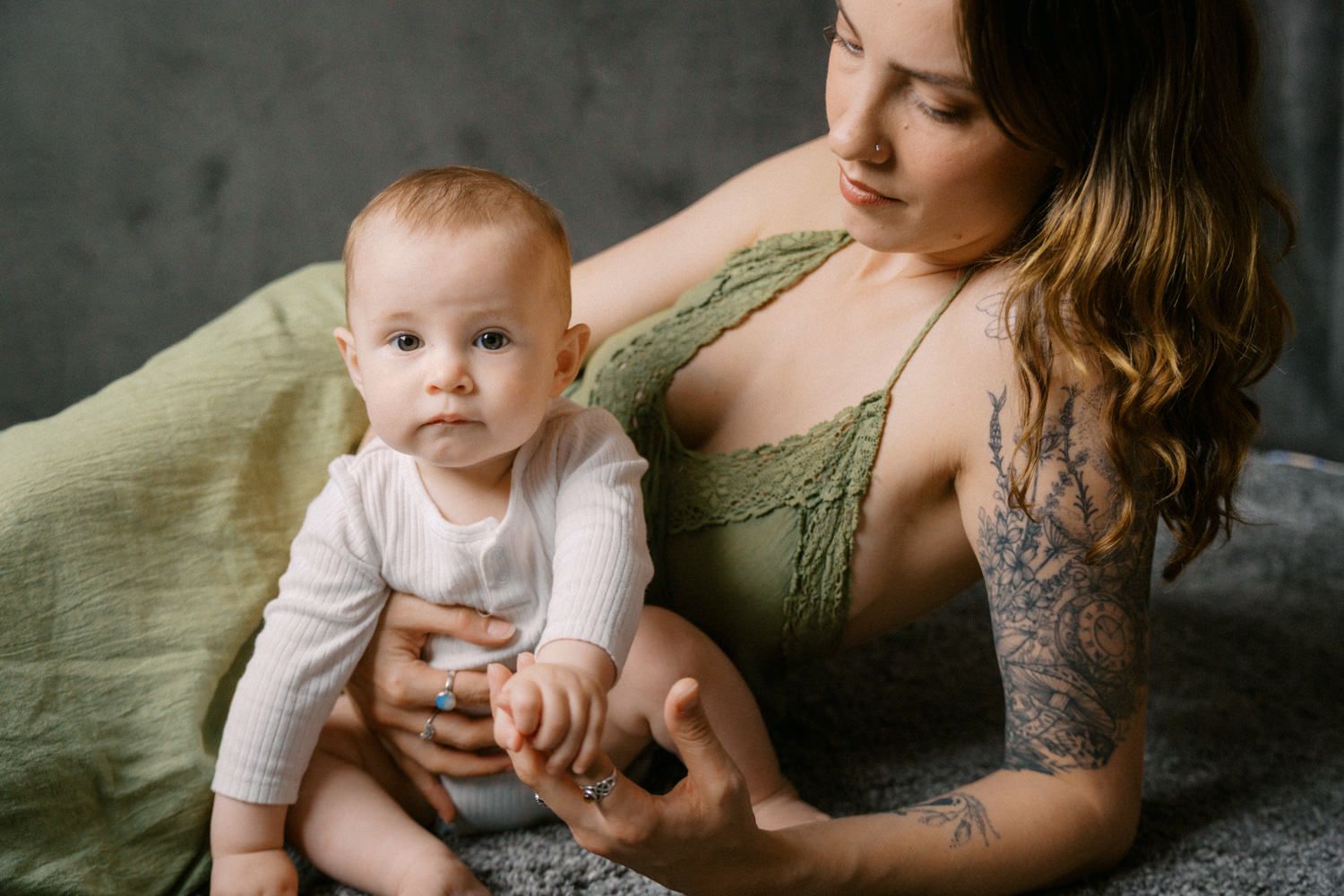 A tattooed mother tenderly holds her baby on a bed with green blankets in a cozy indoor setting.