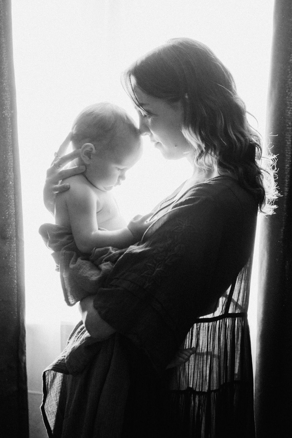Black and white photo of a mother tenderly holding her newborn baby near a bright window.