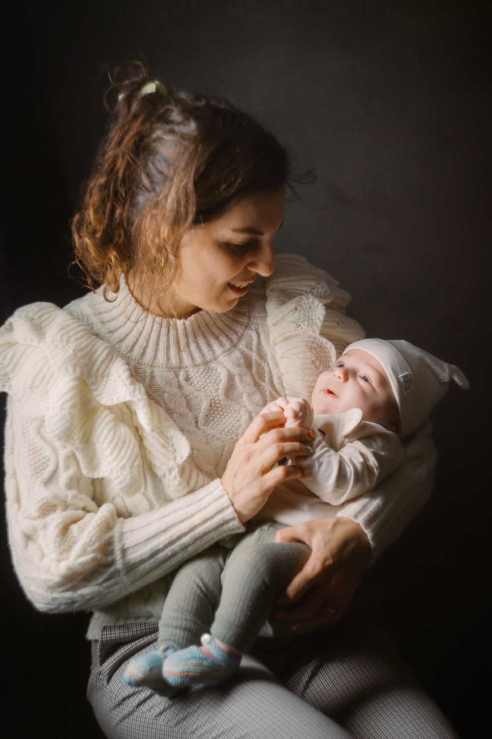 A mother in a white cable-knit sweater tenderly holds her newborn baby against a dark background.