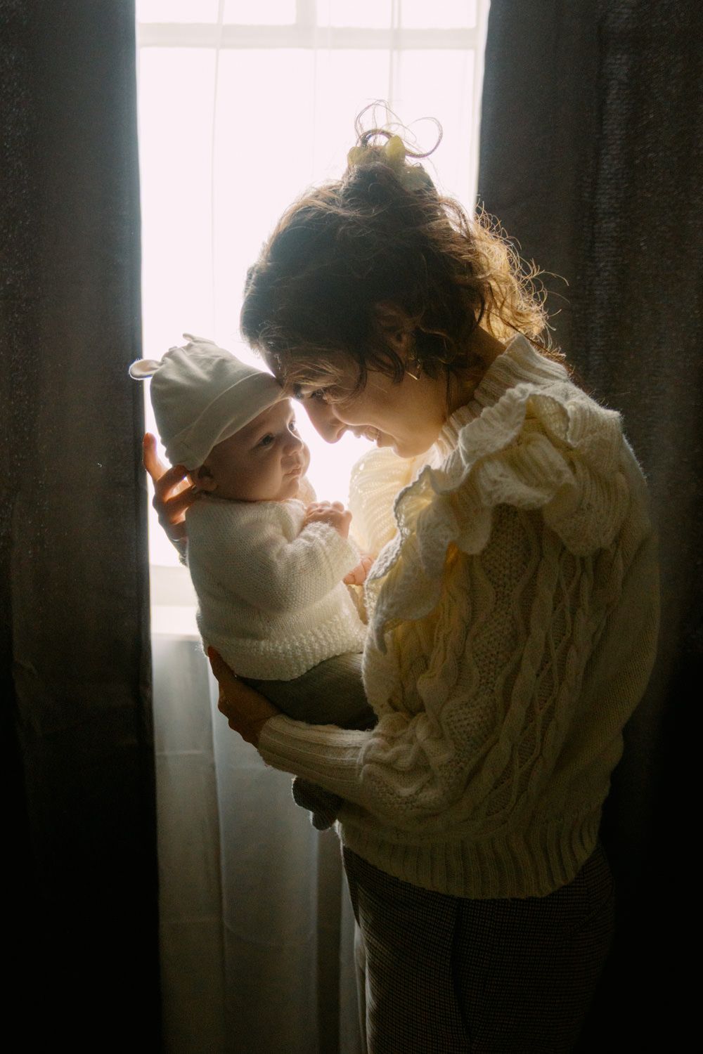 A mother in a white knit sweater tenderly holds her newborn baby near a sunlit window.