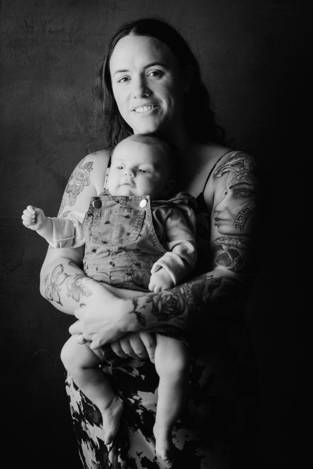 Black and white portrait of tattooed mother holding her baby, smiling warmly in a studio setting.