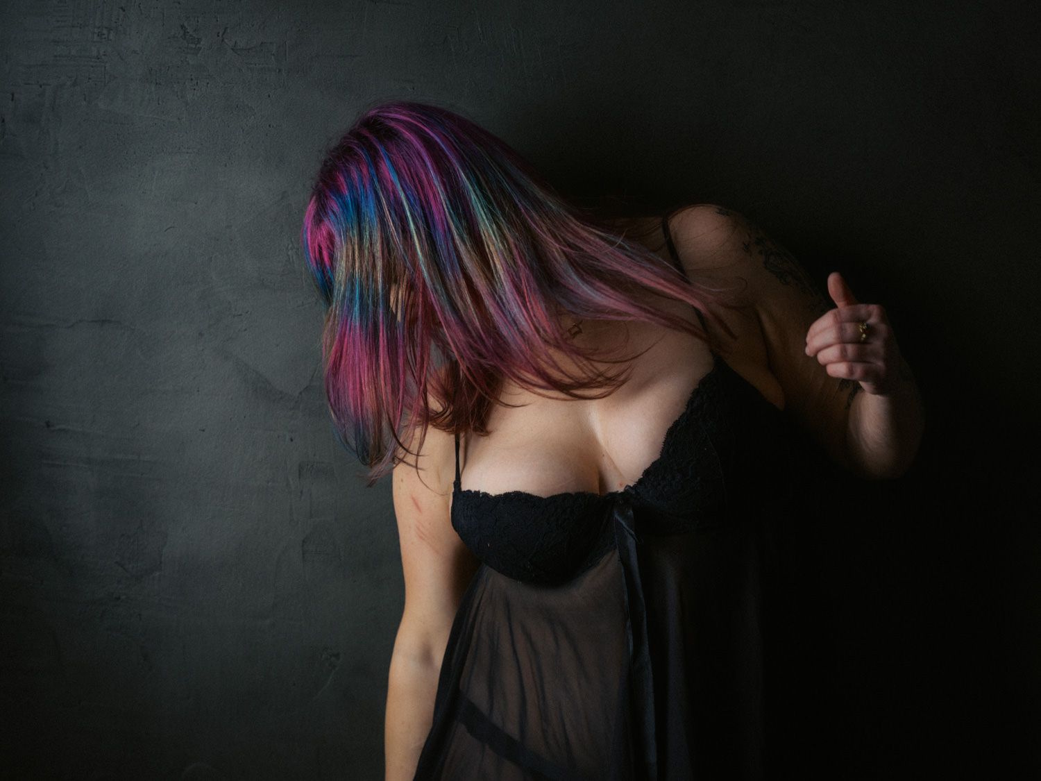 Person with vibrant multicolored hair in black sheer dress posed dramatically against dark wall.