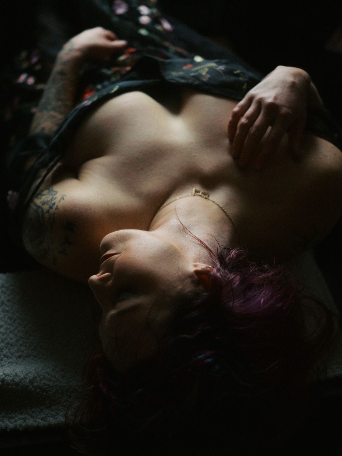 Person with purple hair and tattoos lying down, wearing a floral top, in moody dark lighting.