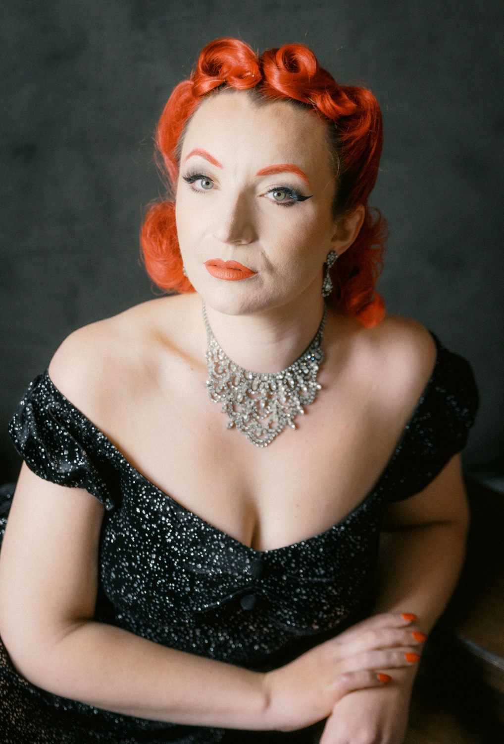 Woman with vintage red pin-up hairstyle wearing black sparkly off-shoulder dress and silver statement necklace poses elegantly.