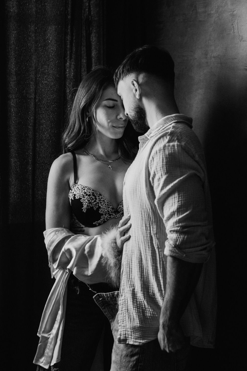 Black and white photo of a romantic couple embracing intimately in a dark studio setting, woman in lace bra and open shirt.