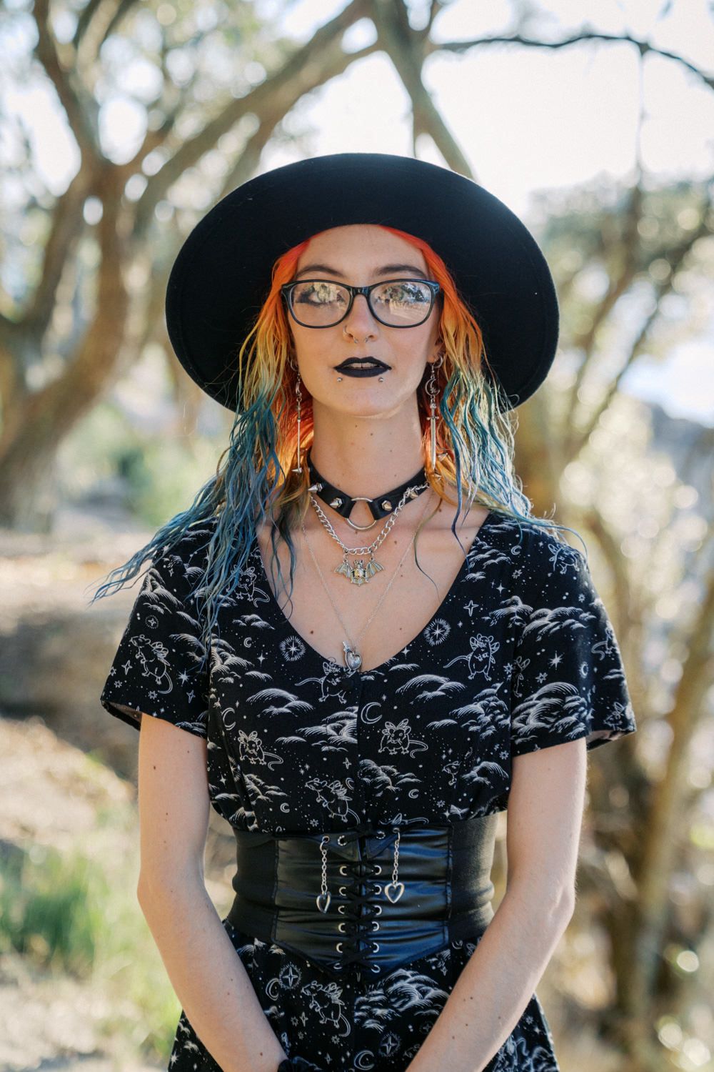 Woman with colorful hair, black hat, glasses, and dark lipstick wearing gothic fashion outdoors.