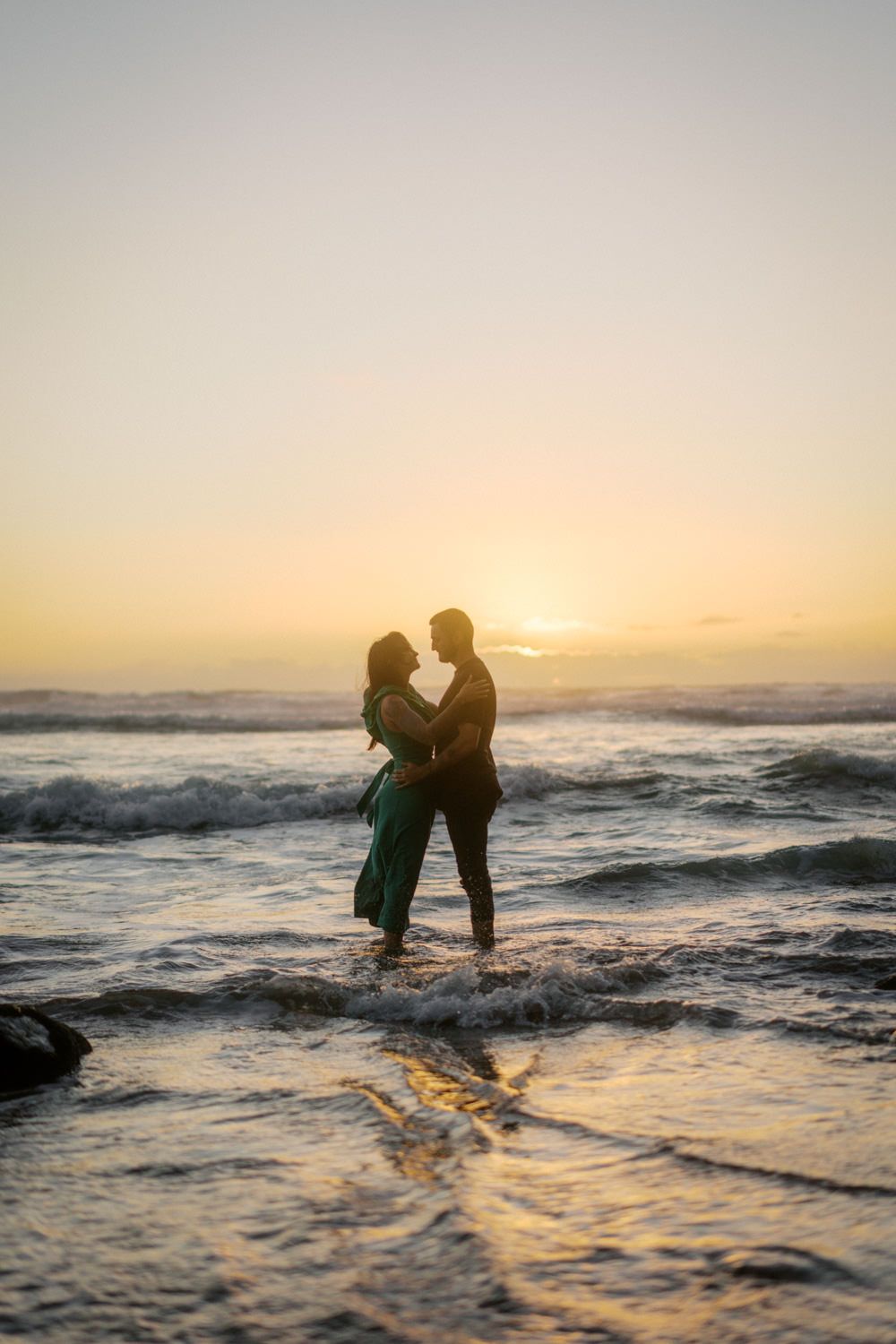 Romantic couple silhouetted against a golden sunset, standing in ocean waves on a beach, woman in green dress.