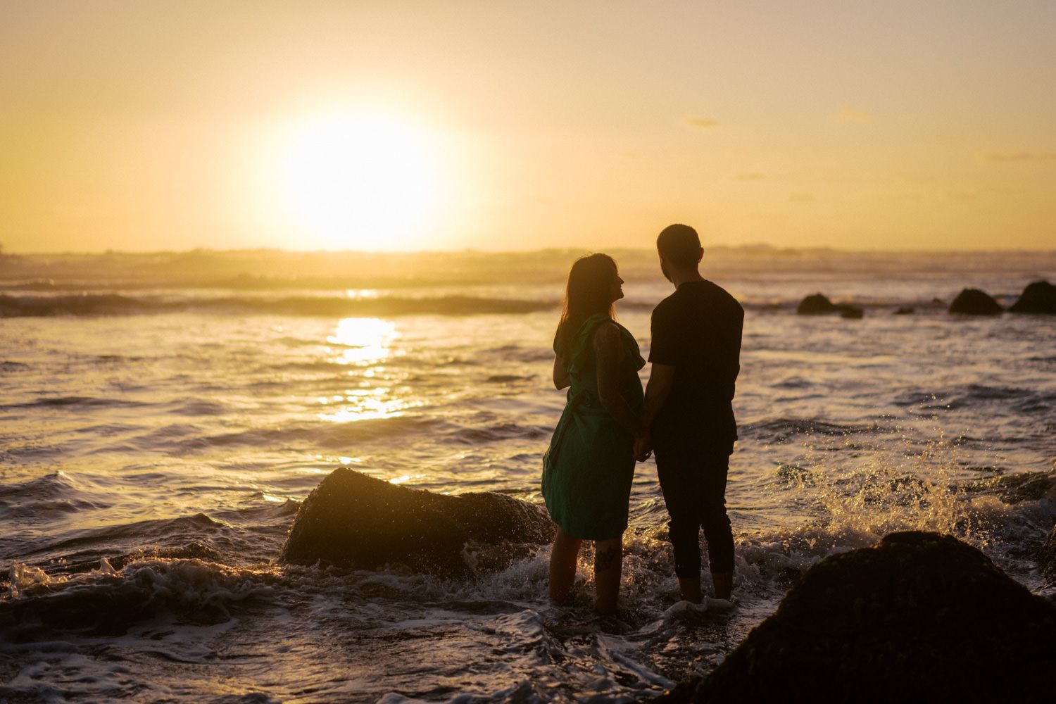 Couple silhouetted against golden sunset standing on rocky ocean shore with waves splashing around their feet.
