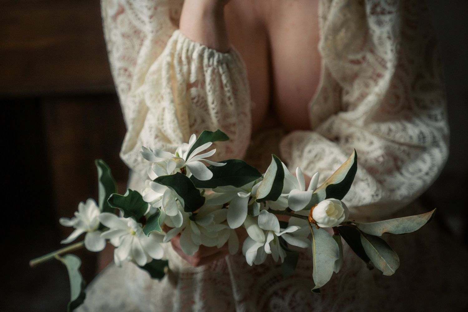 Person in lace dress holding delicate white floral branch with green leaves in a softly lit, moody setting.
