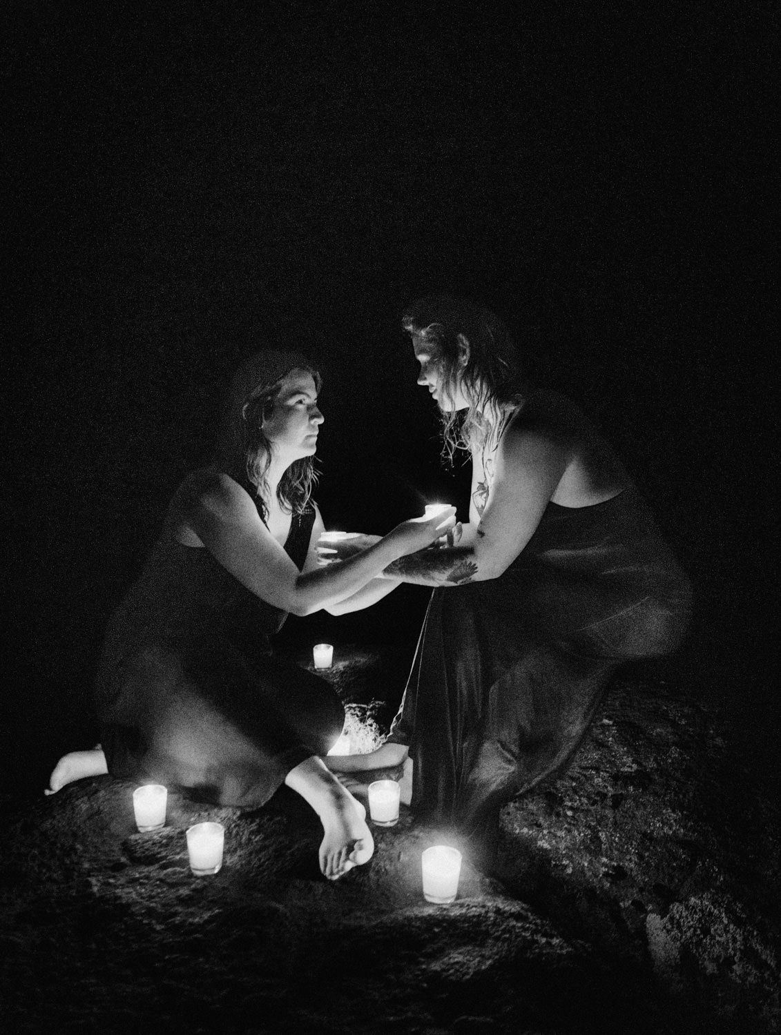 Two women kneel together by candlelight in a dark, intimate black and white photo with glowing candles surrounding them.