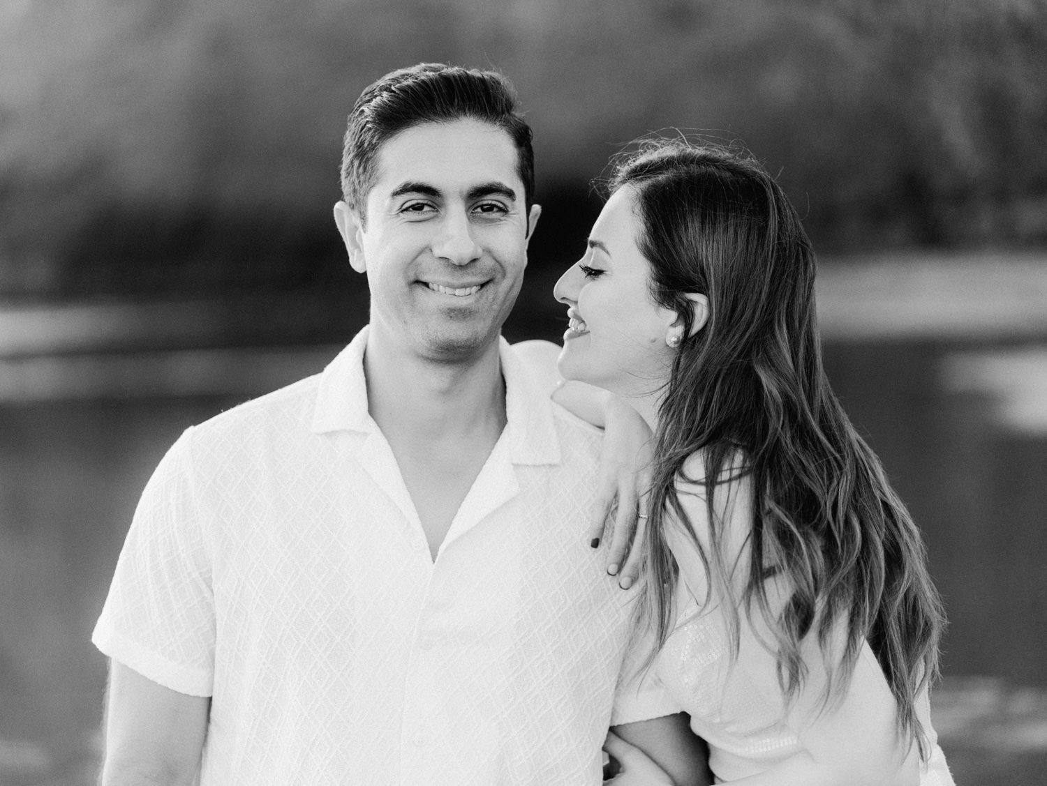 Black and white photo of a smiling couple embracing outdoors near water, woman leaning on mans shoulder.