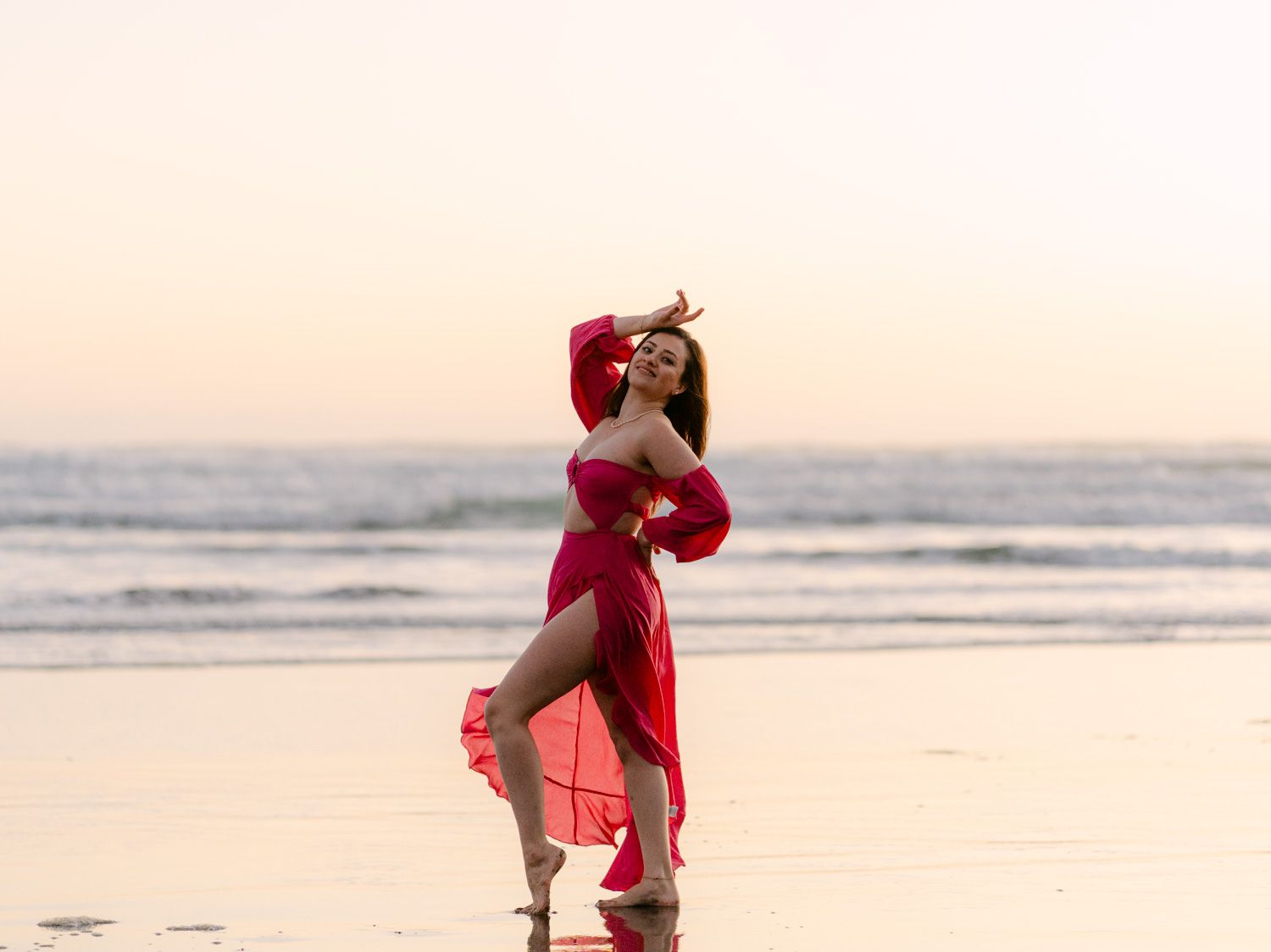 Woman in red dress posing gracefully on a beach at sunset with ocean waves in the background.