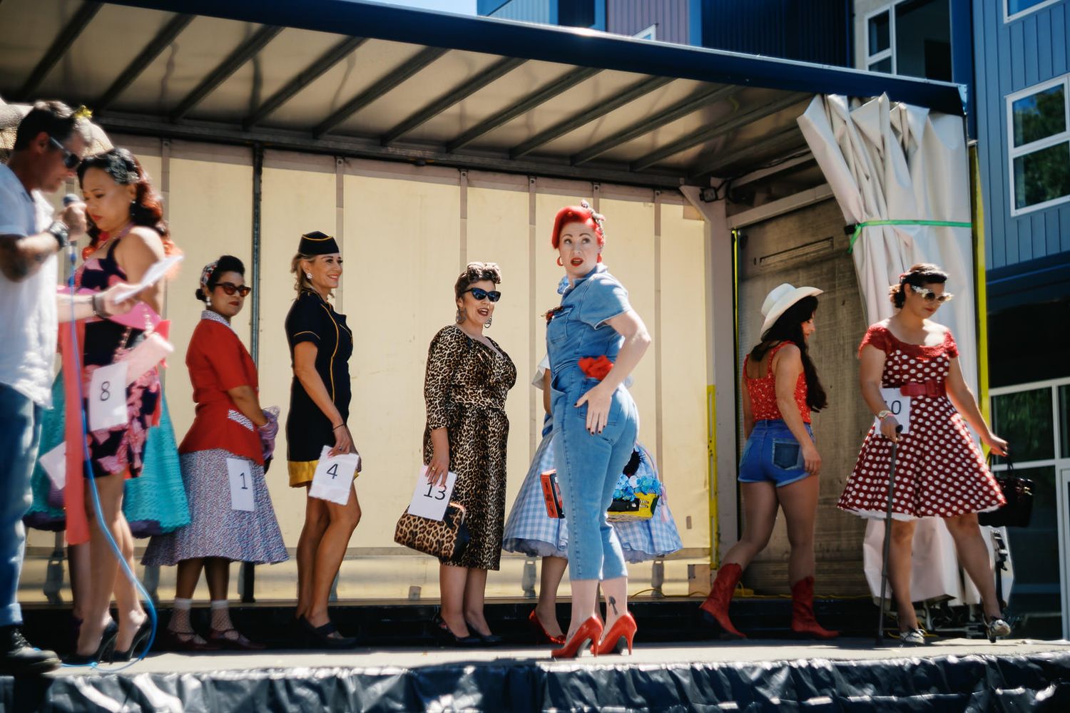 Women in vintage 1950s pin-up style dresses pose on a stage during a retro fashion show outdoors.