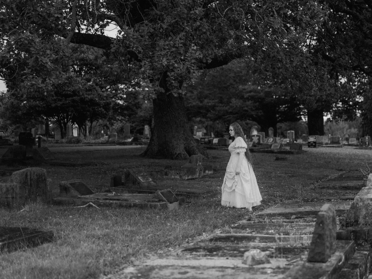 Woman in white dress standing in historic cemetery beside large tree, black and white photography.