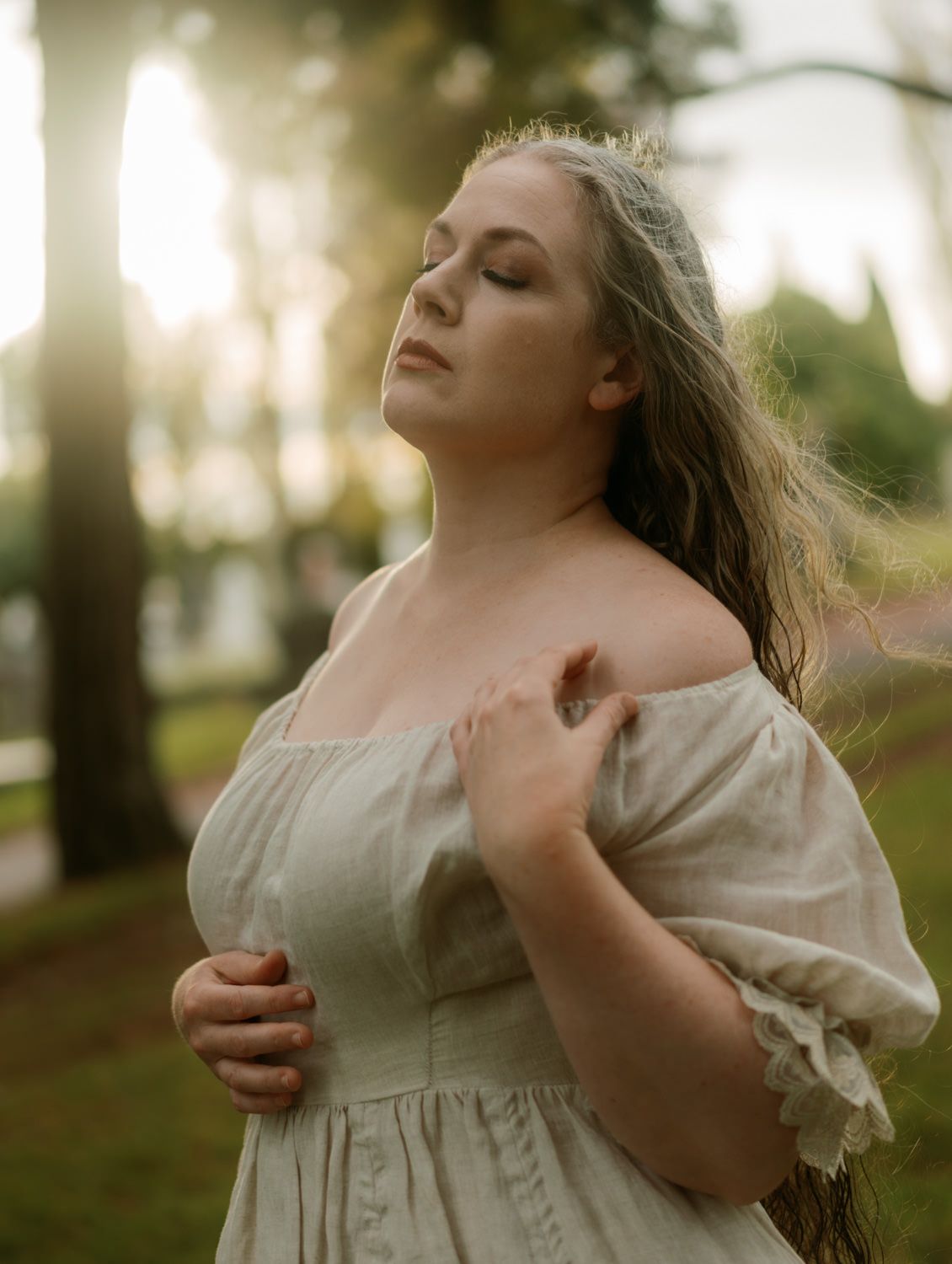Woman in off-shoulder linen dress with long hair poses outdoors in golden sunset light.