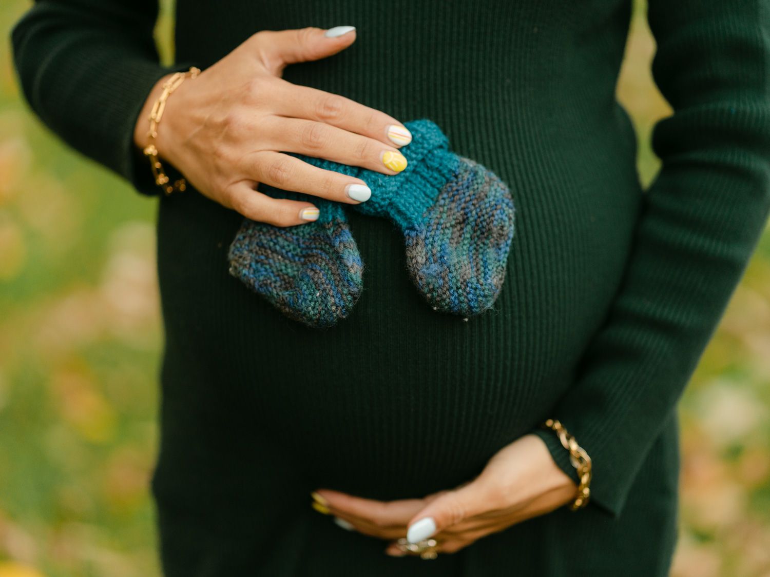 Pregnant woman in green sweater holding tiny knitted baby booties against her baby bump outdoors.