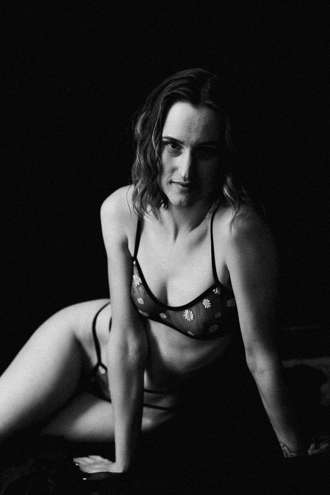 Black and white boudoir photo of a woman in sheer floral lingerie posing confidently against a dark background.