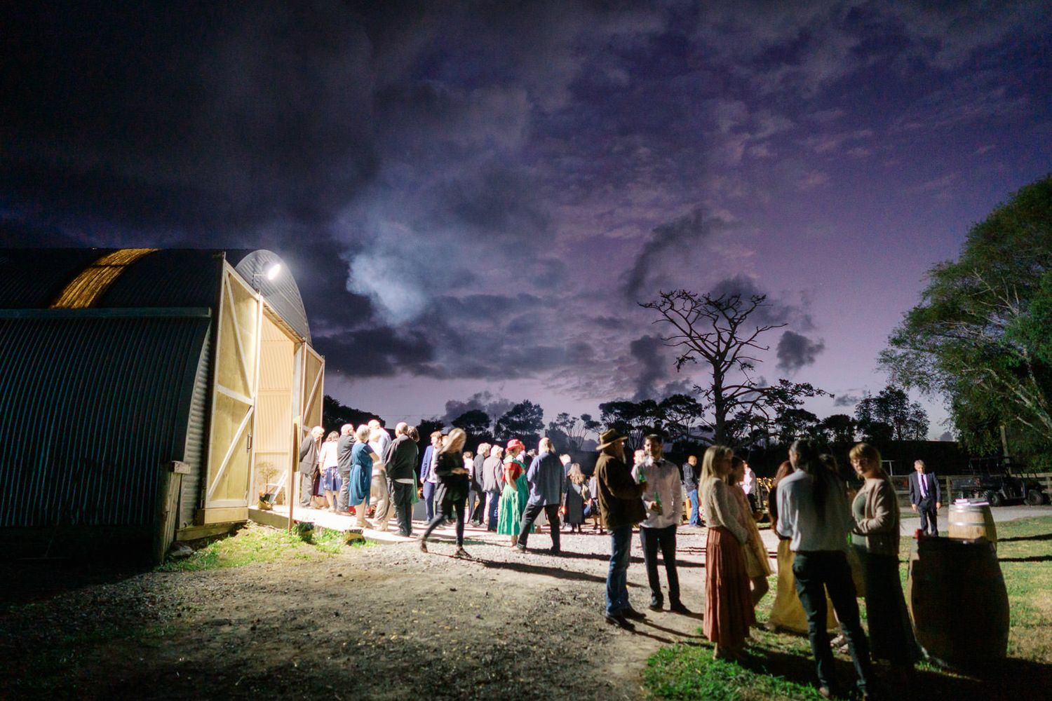 Crowd queuing outside a barn venue at night under a dramatic purple sky with trees silhouetted in background.