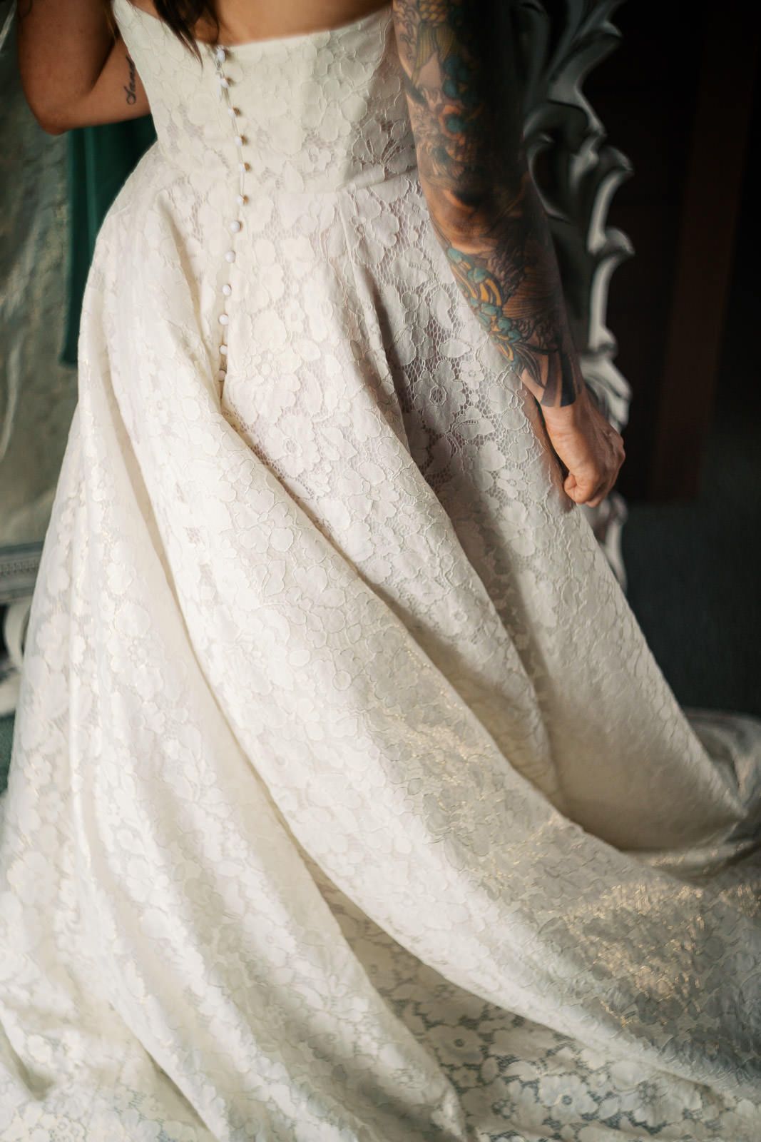 Bride in a lace wedding gown with button back, held by tattooed hands adjusting the flowing skirt.