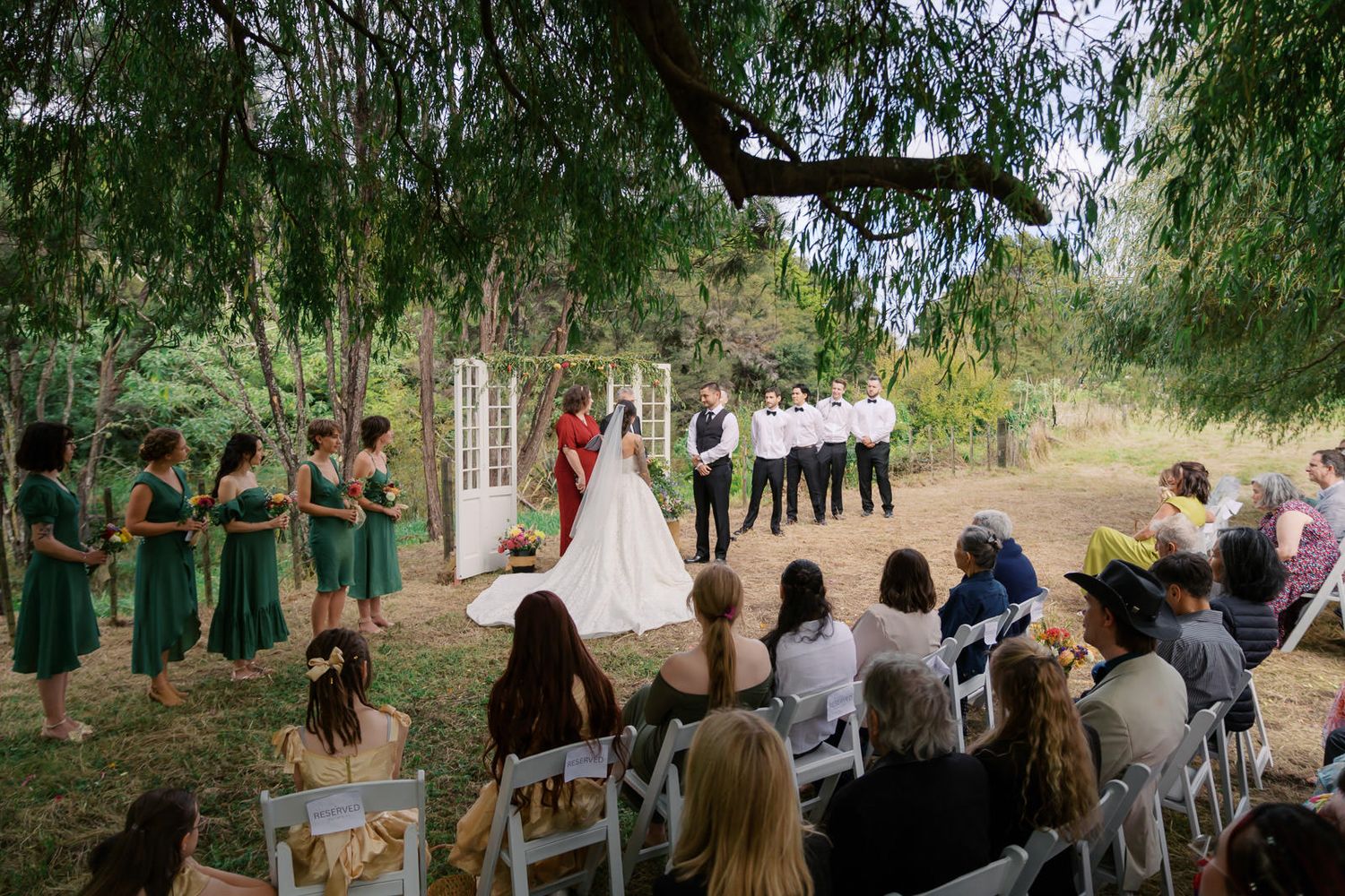 Outdoor wedding ceremony under willow trees with bridesmaids in green dresses and seated guests.