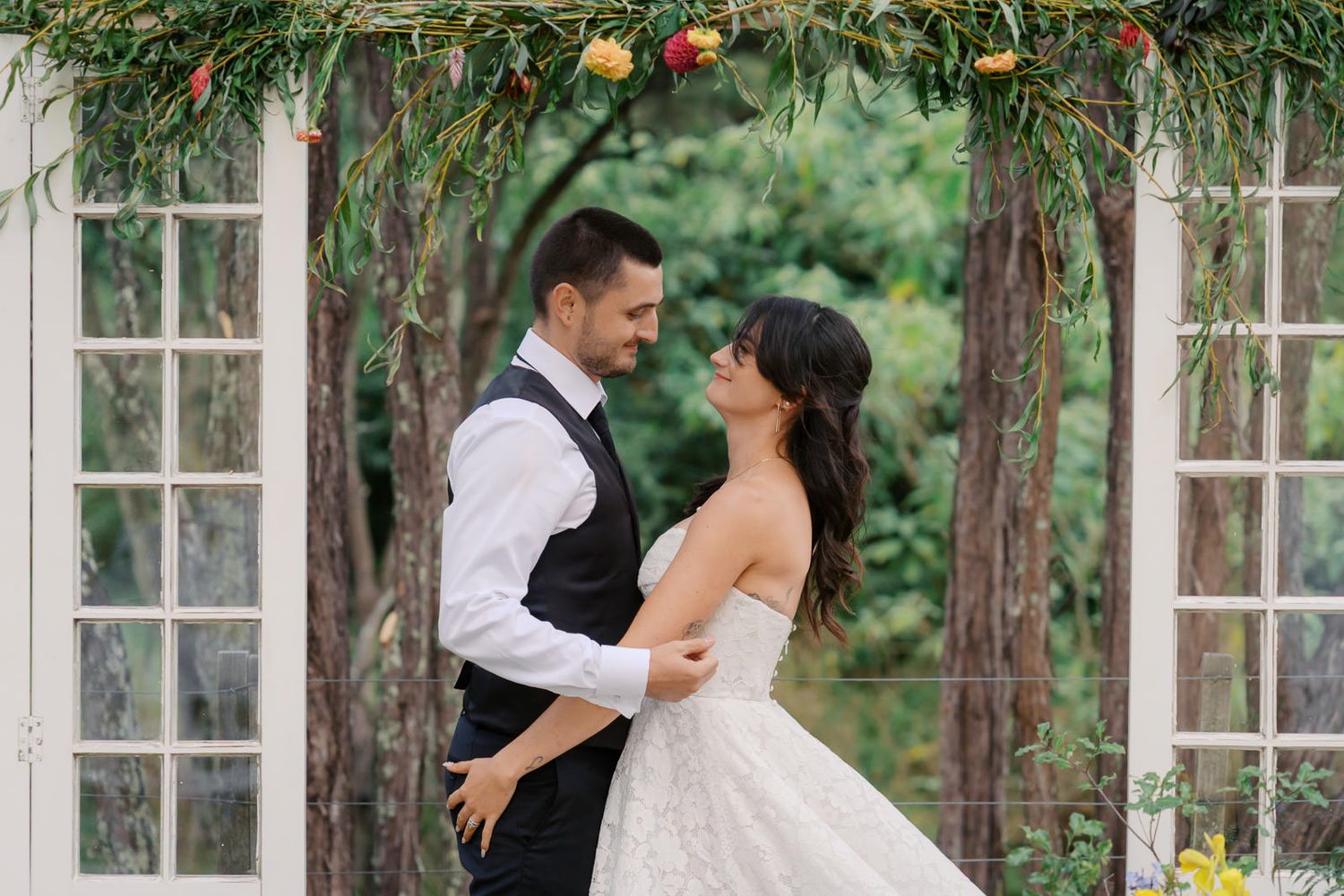 A bride and groom share a romantic moment under a floral arch at an outdoor wedding ceremony.