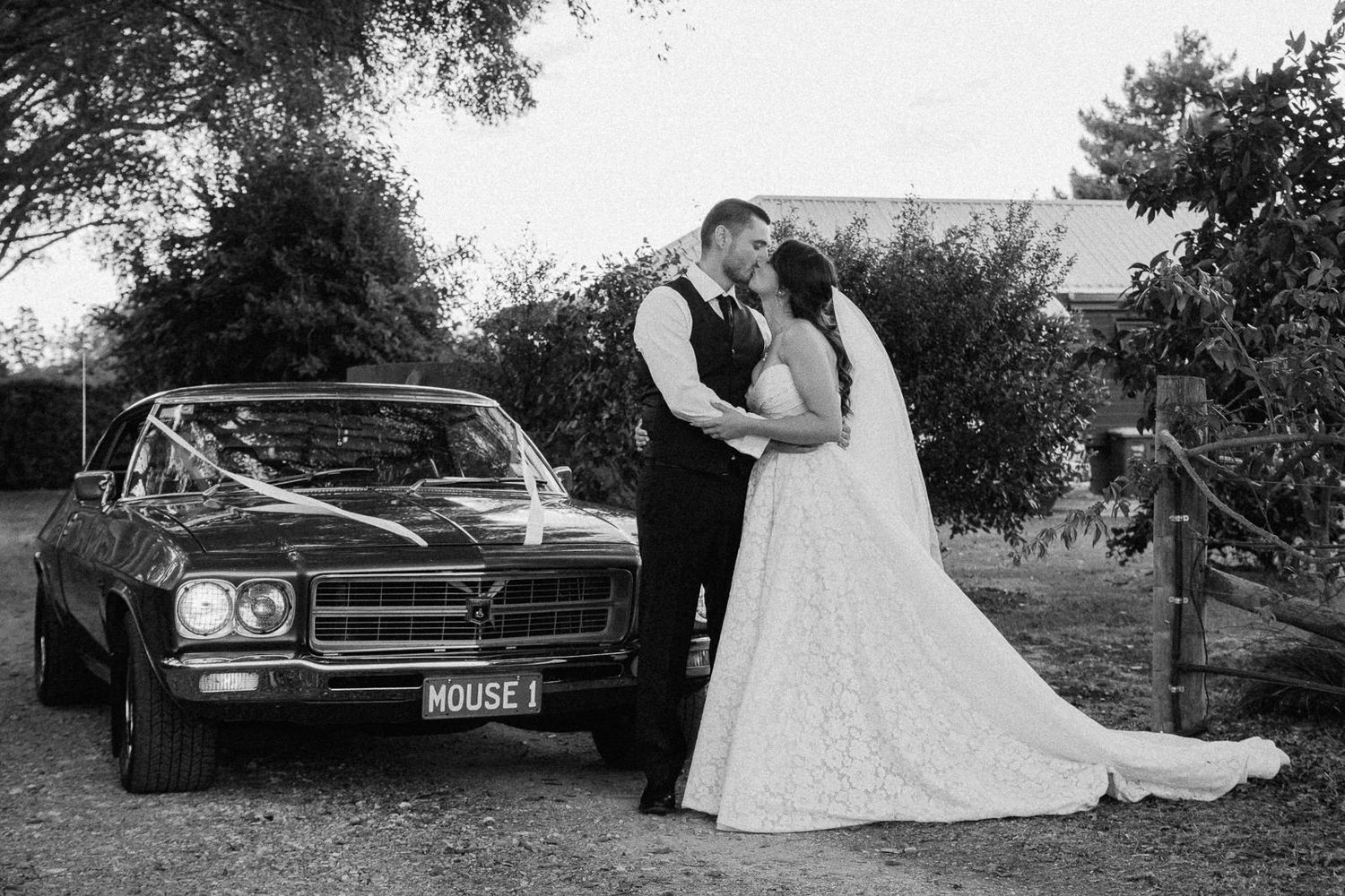 Black and white wedding photo of bride and groom kissing beside a classic car with ribbon decorations outdoors.