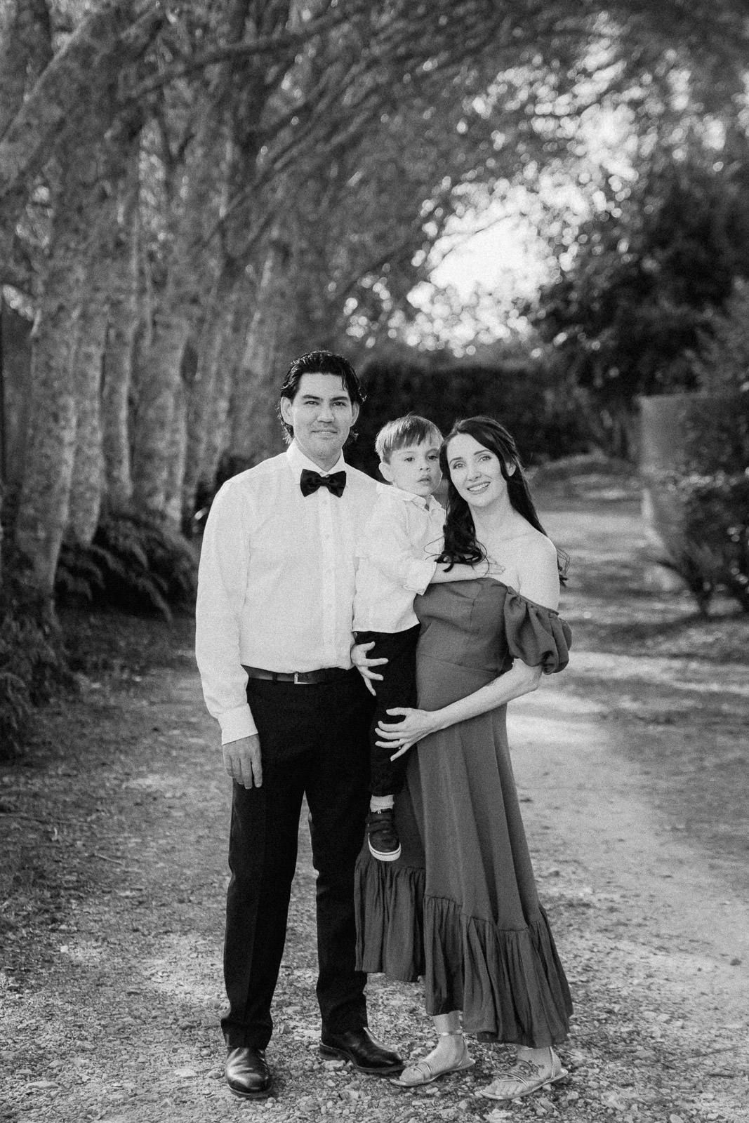 Black and white family portrait of parents and toddler on a tree-lined path, mother in off-shoulder dress showing baby bump.