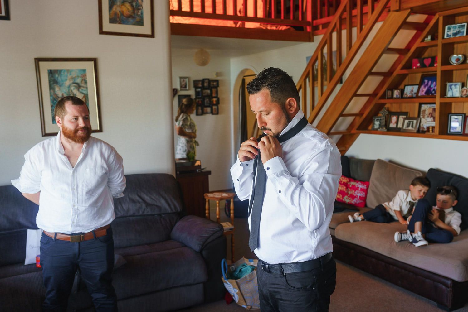 Groom adjusting his tie in a living room while groomsmen prepare nearby before a wedding ceremony.