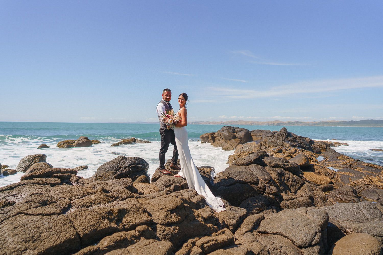 Bride and groom stand on rocky coastal shore with ocean waves and blue sky backdrop on their wedding day.