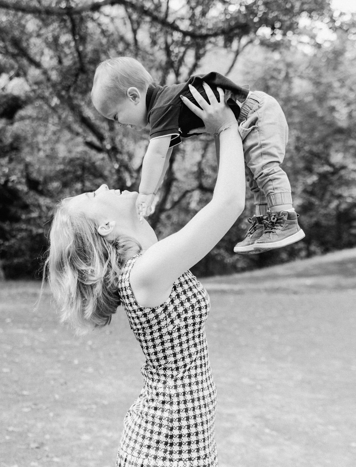Black and white photo of a smiling mother lifting her baby boy in the air outdoors.