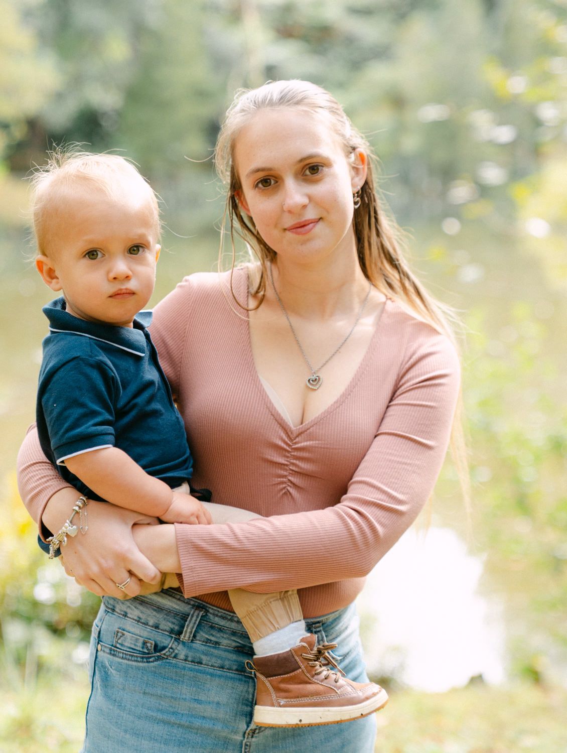 Young blonde woman in pink top holds a toddler boy outdoors near a pond with lush green trees.