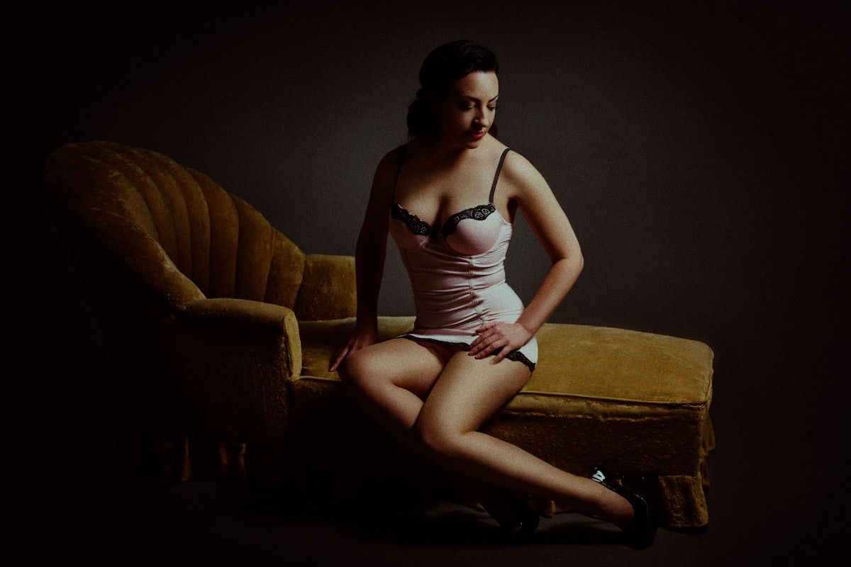 A woman in lingerie poses gracefully beside a vintage gold velvet sofa in dramatic low-key lighting.
