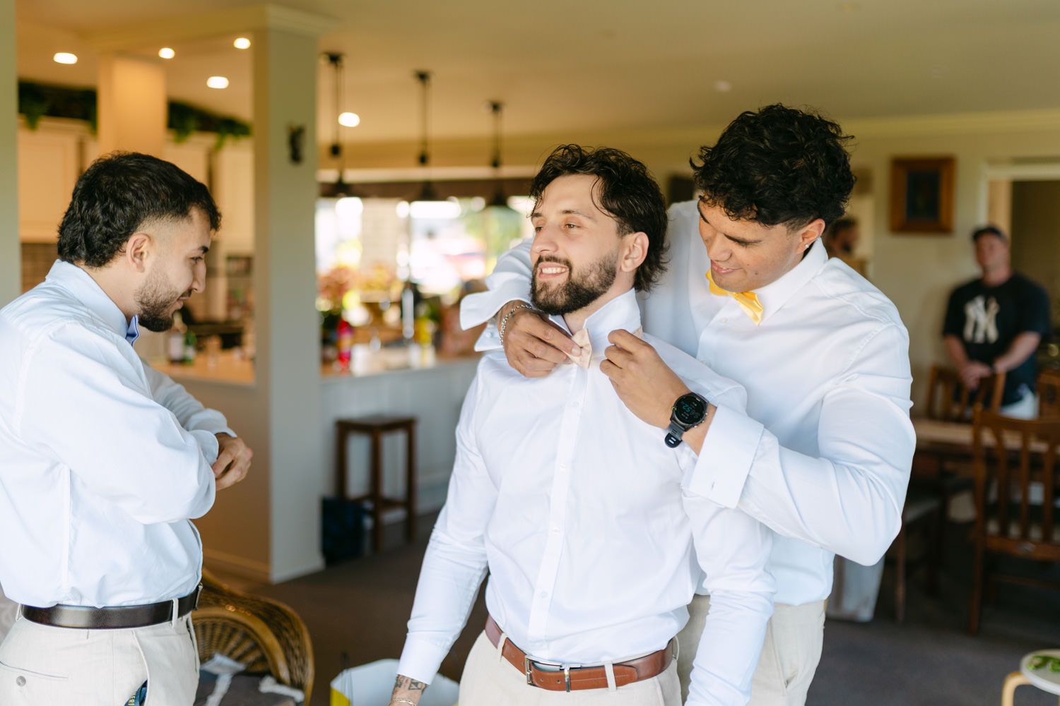 Groomsmen helping groom adjust bow tie while getting ready indoors before wedding ceremony.