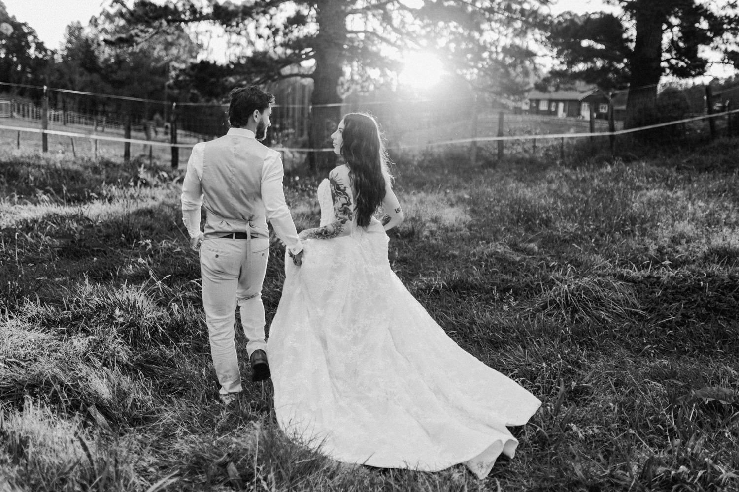 Black and white photo of a bride and groom walking hand in hand through a sunlit field on their wedding day.