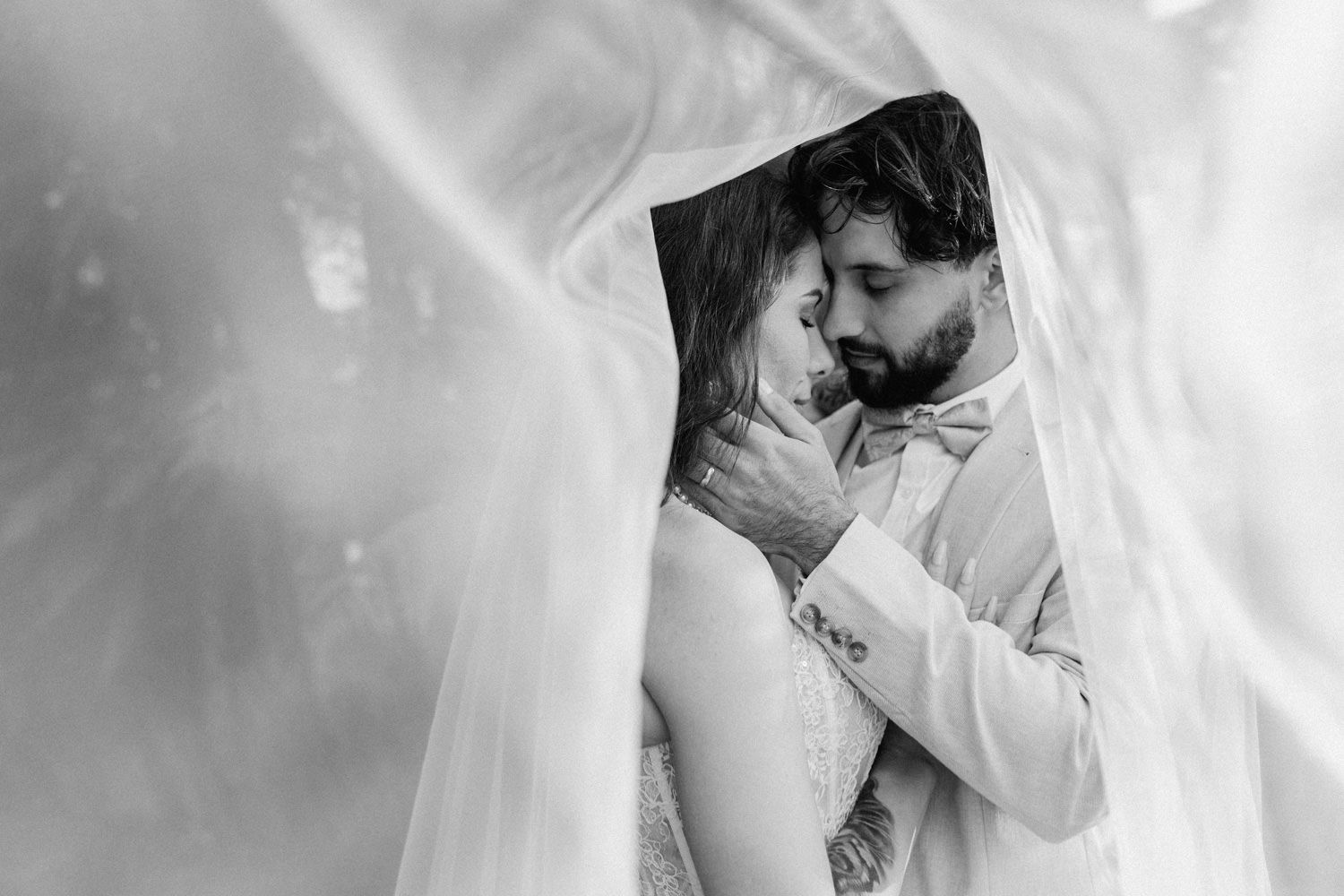 Black and white wedding photo of bride and groom embracing tenderly under a flowing veil.