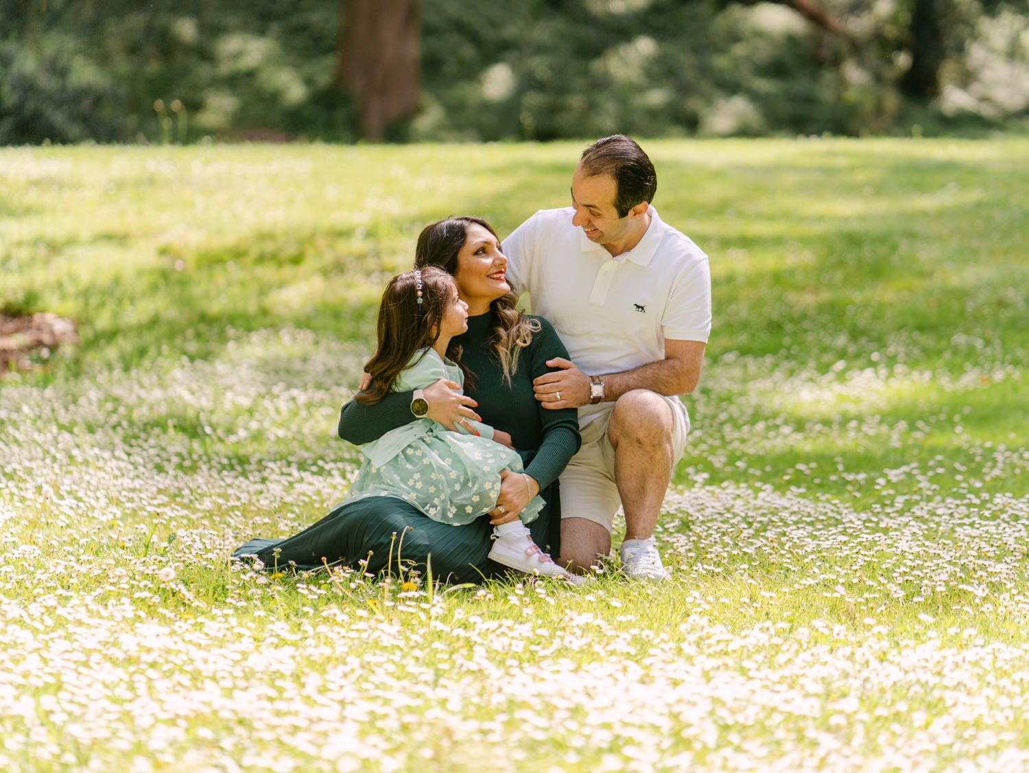 Family of three sitting together in a sunny meadow filled with white wildflowers, sharing a joyful moment outdoors.