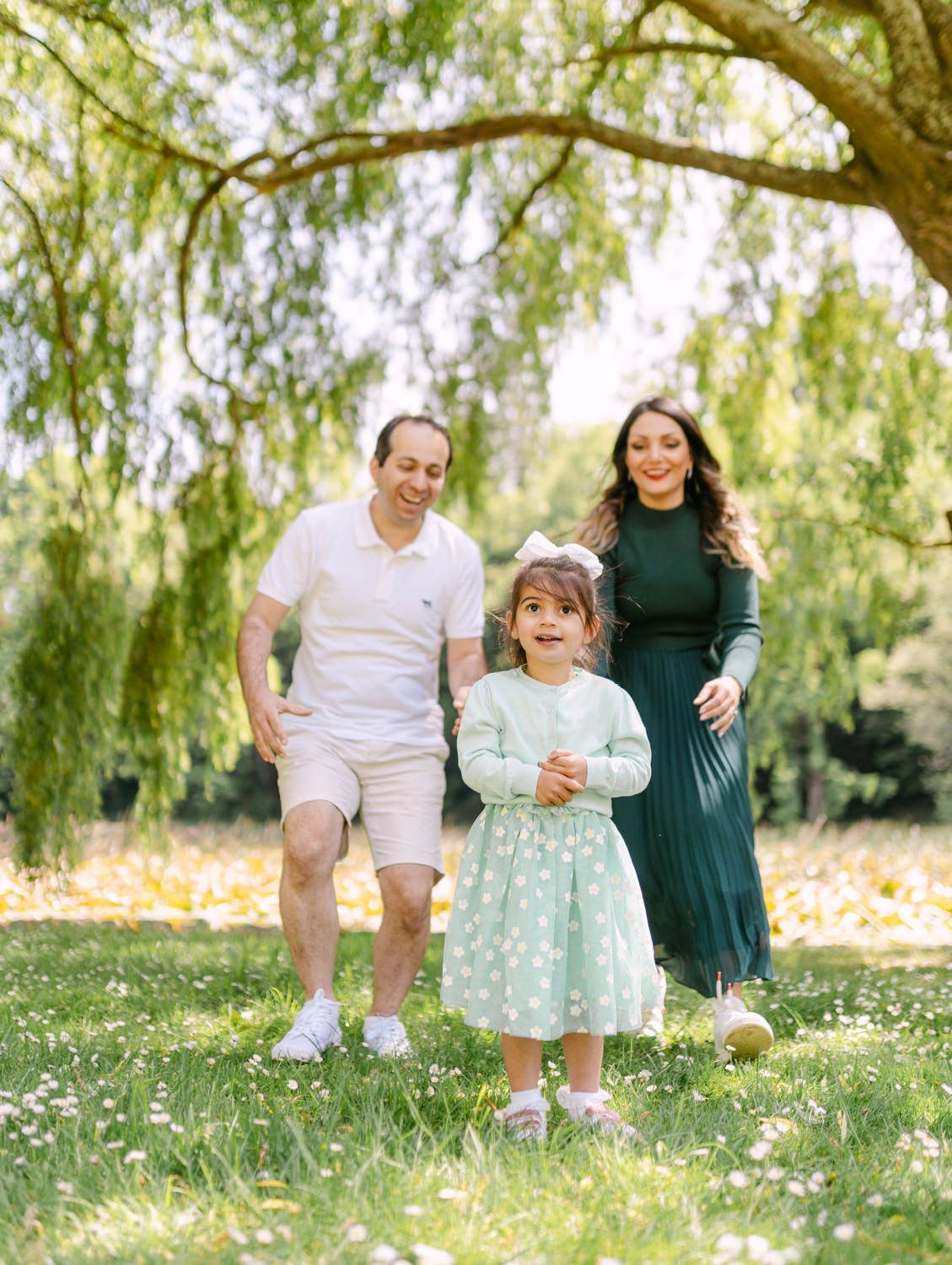 A happy family of three walks in a sunny park, with a little girl in a mint dress between her smiling parents.