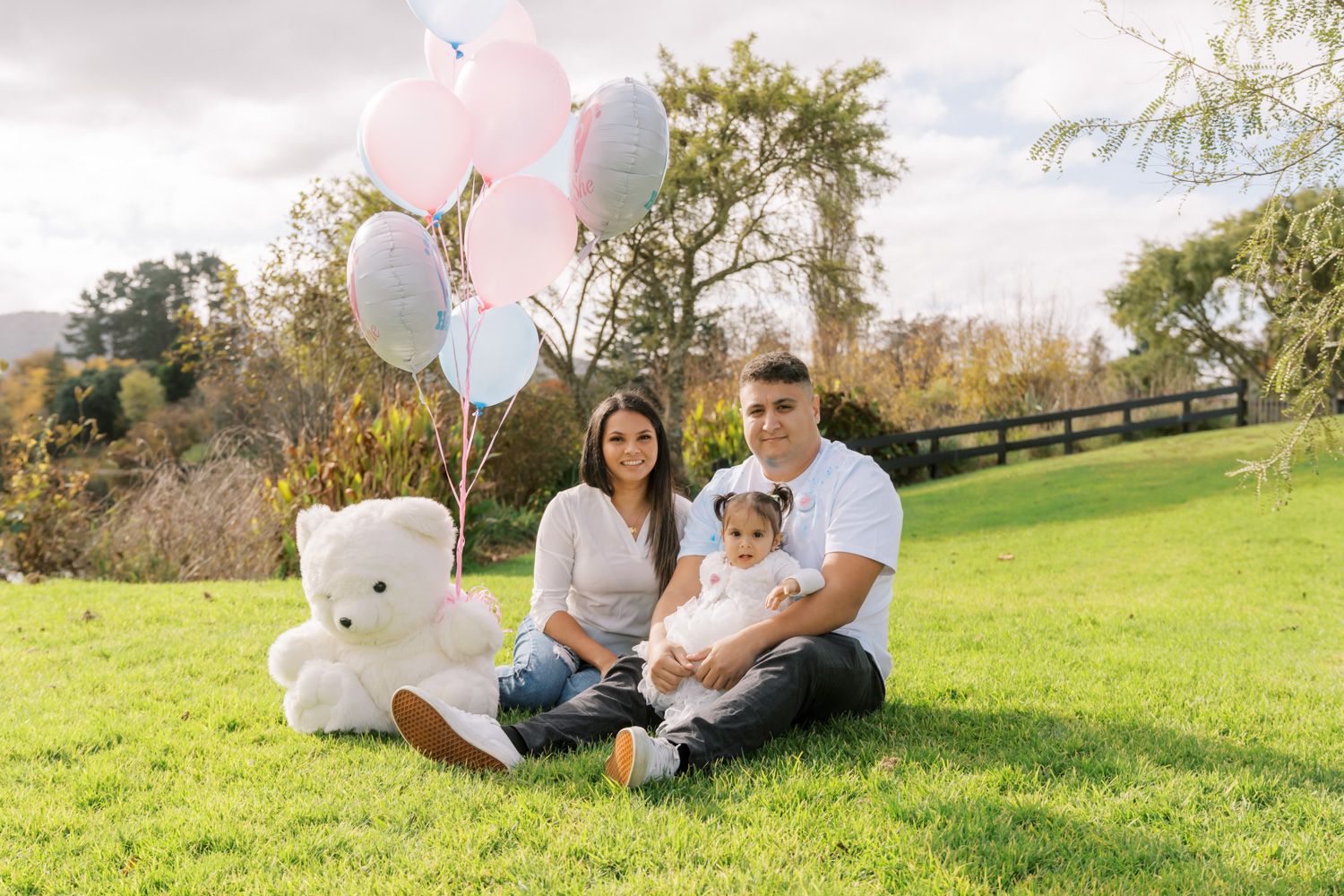 Family of three sitting on grass with pink and blue balloons and white teddy bear for gender reveal.