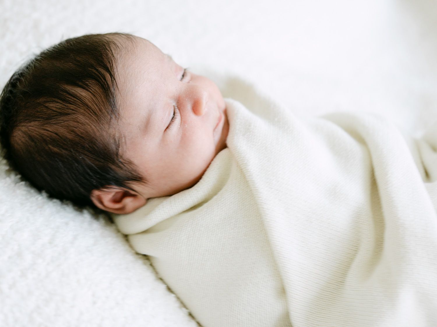Peaceful newborn baby sleeping swaddled in a cream white blanket on a soft white surface.