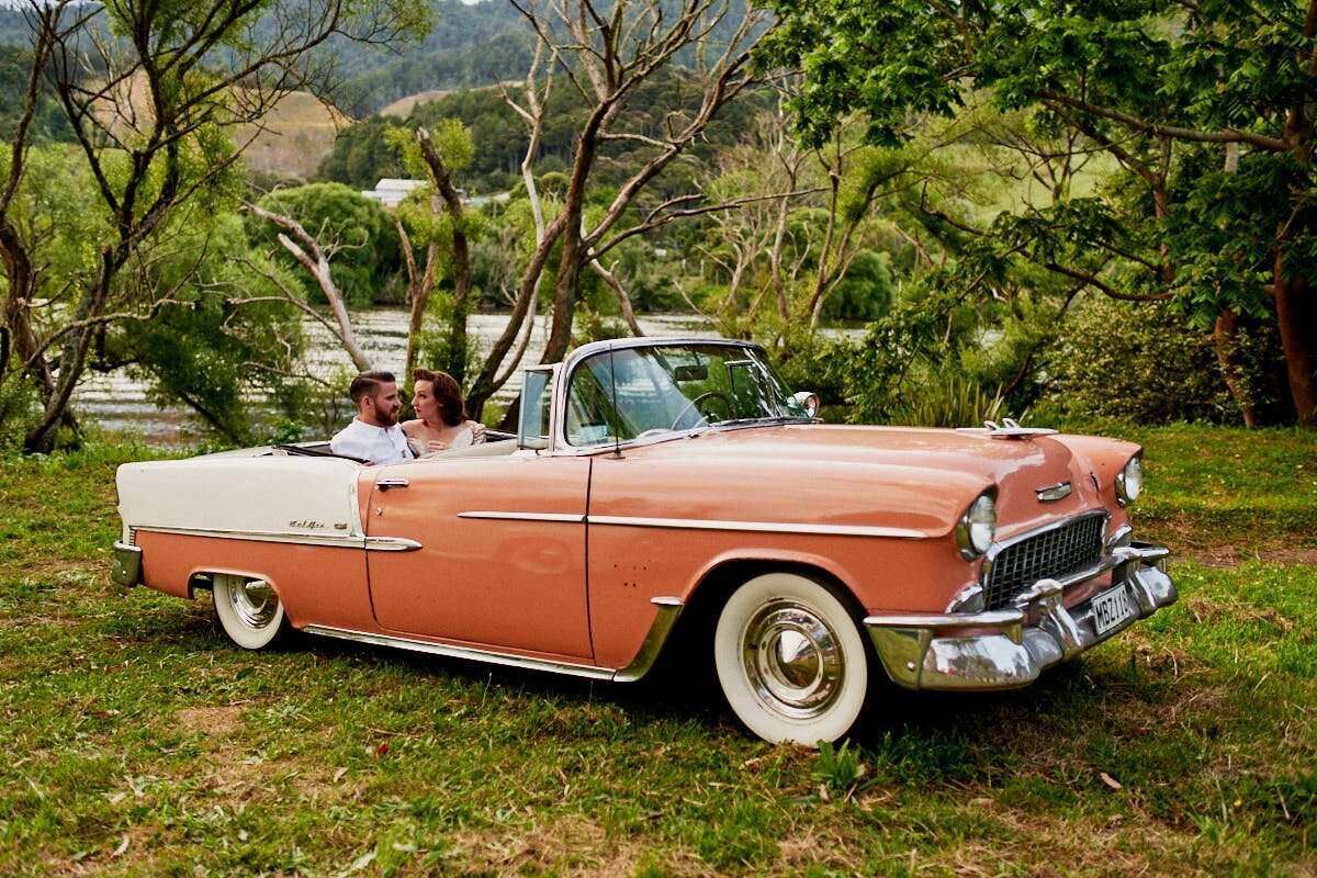 Couple sitting in a vintage pink 1955 Chevrolet Bel Air convertible on green grass with lush trees behind.