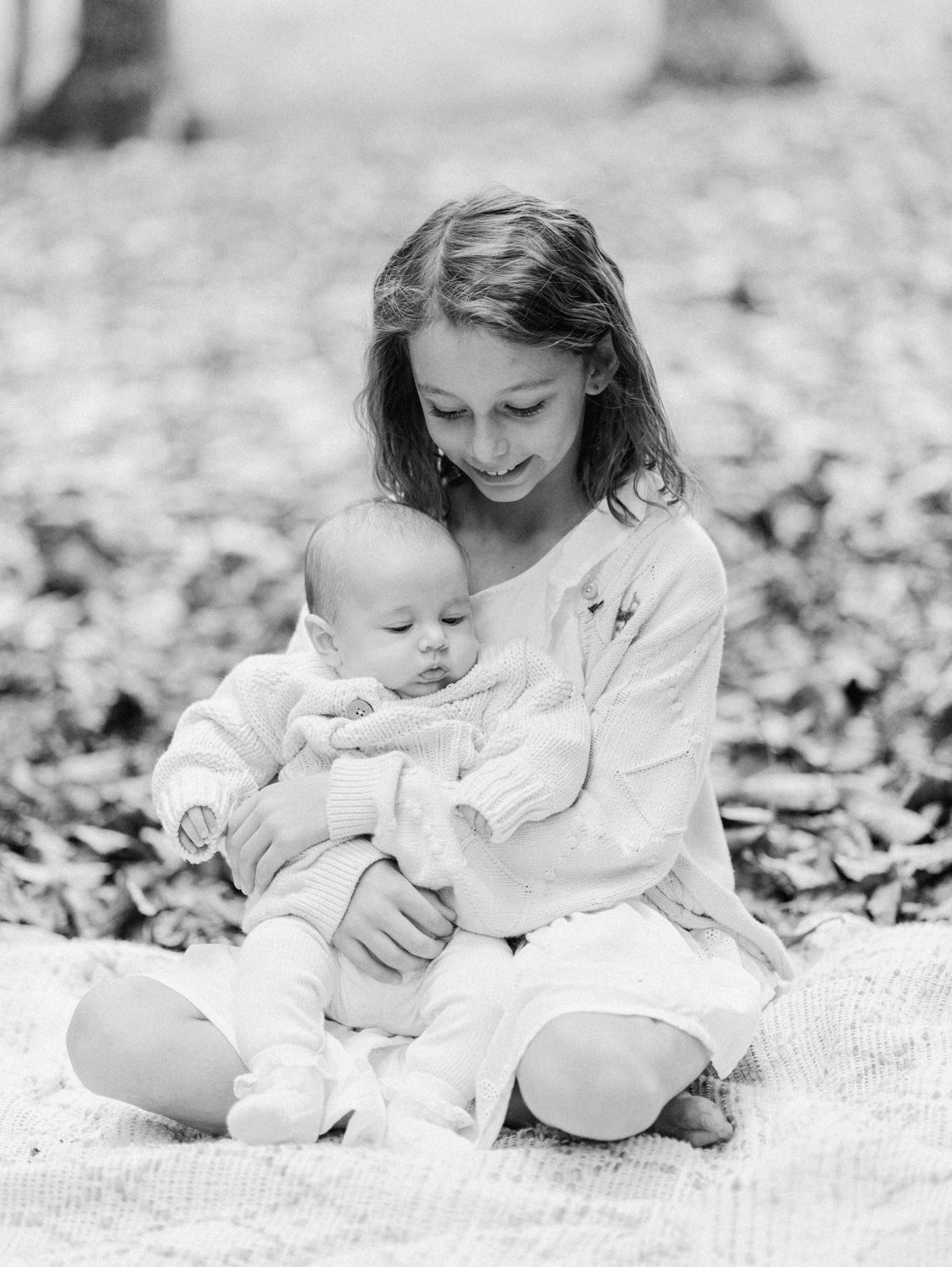 Black and white photo of a young girl tenderly holding a sleeping baby on a blanket outdoors.