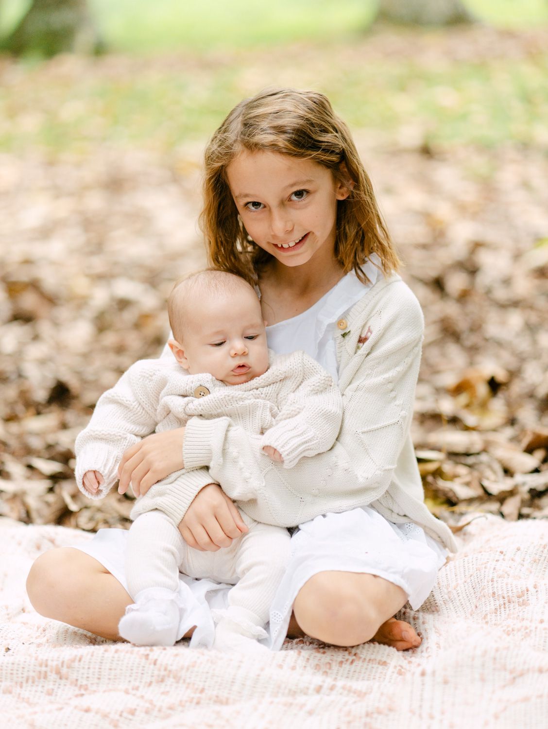 A smiling girl sits on a blanket holding a baby in cream outfits among autumn leaves outdoors.