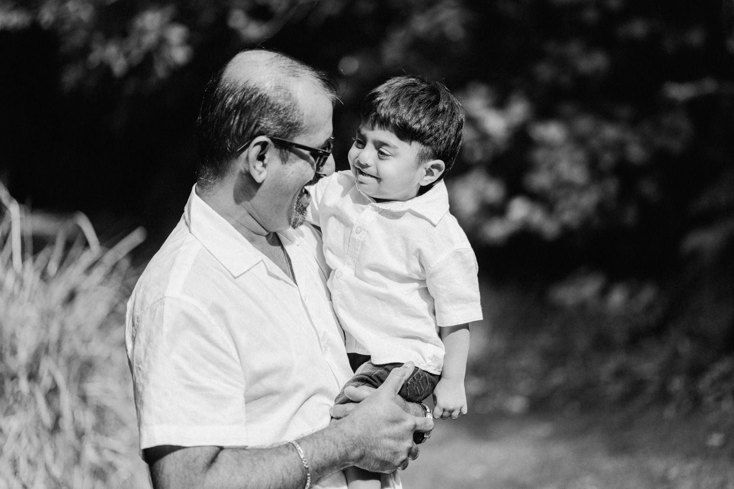 Black and white photo of a grandfather holding and smiling at a young boy outdoors near trees.