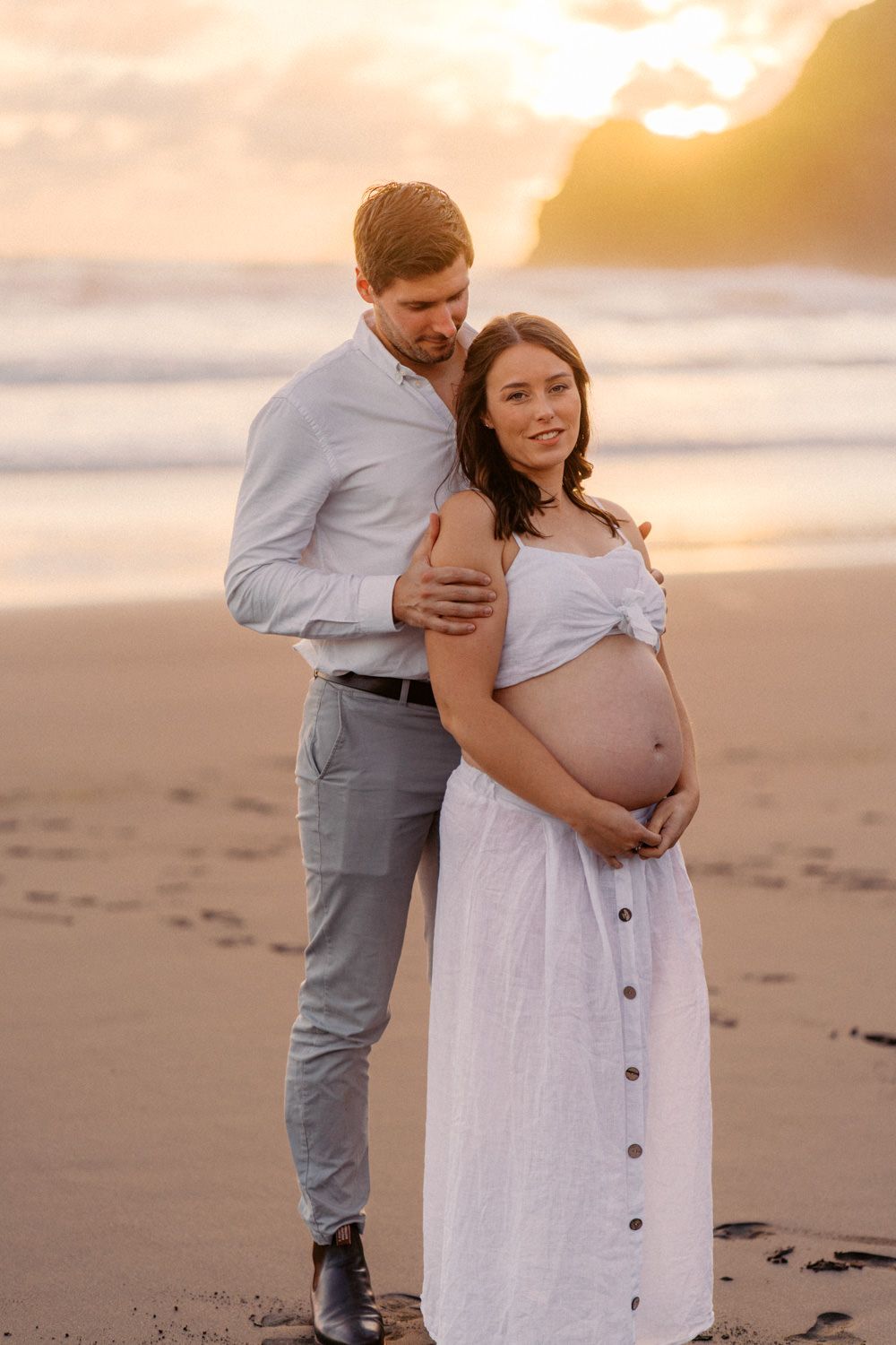 Couple embracing on beach at sunset, pregnant woman in white dress cradling baby bump, romantic maternity photo.