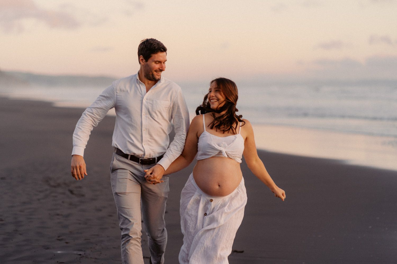 Happy pregnant couple holding hands walking on beach at sunset, woman in white dress showing baby bump.