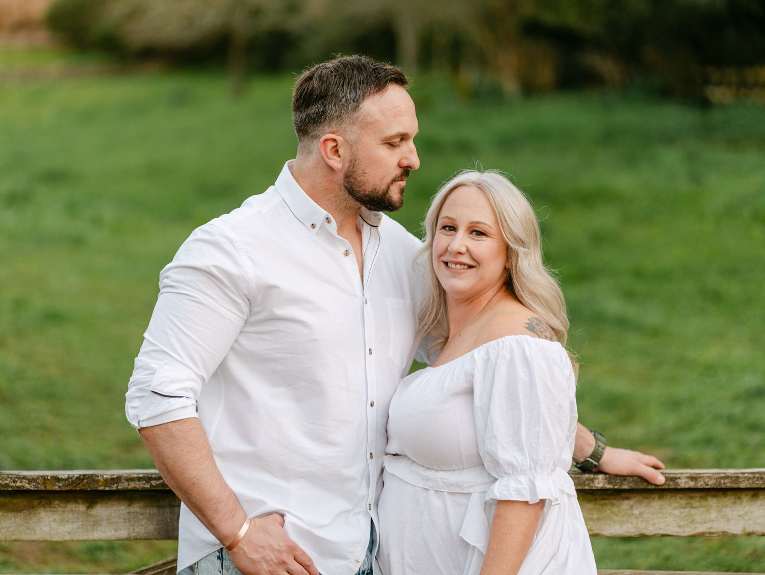 Couple in matching white outfits posing outdoors near a wooden fence with lush green grass background.