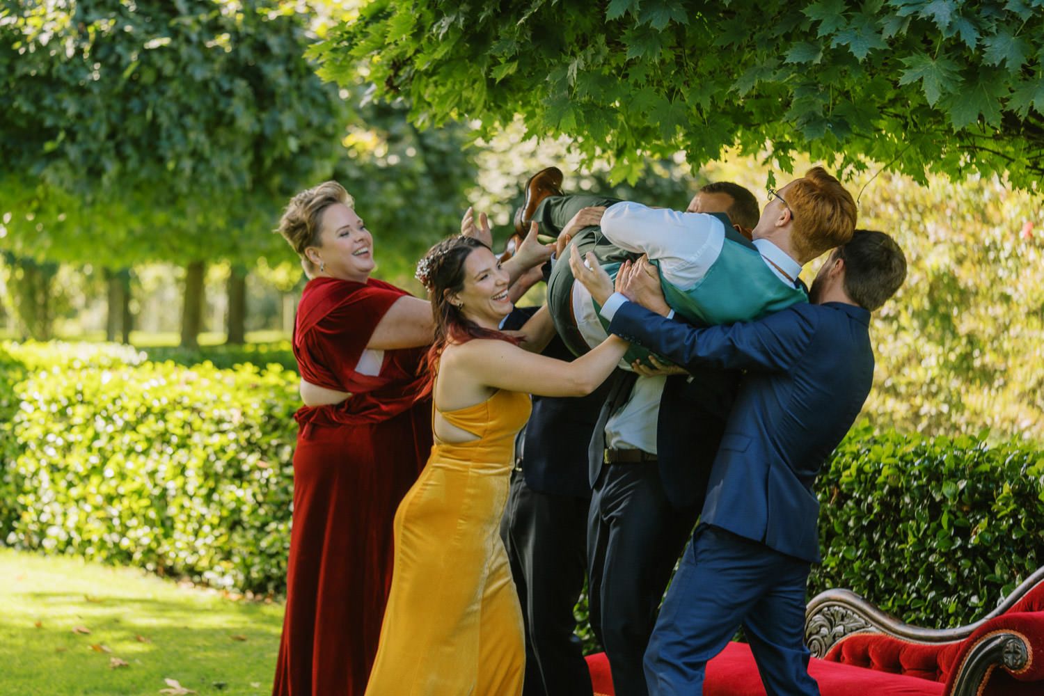 Wedding guests in formal attire joyfully lifting a person outdoors in a lush green garden setting.